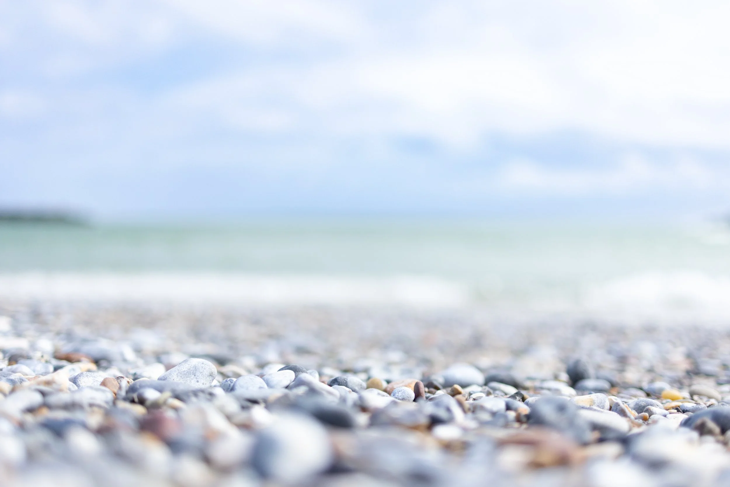 Blick auf einen Kiesstrand mit kleinen Steinen und im Hintergrund das Meer und einen bewölkten Himmel.