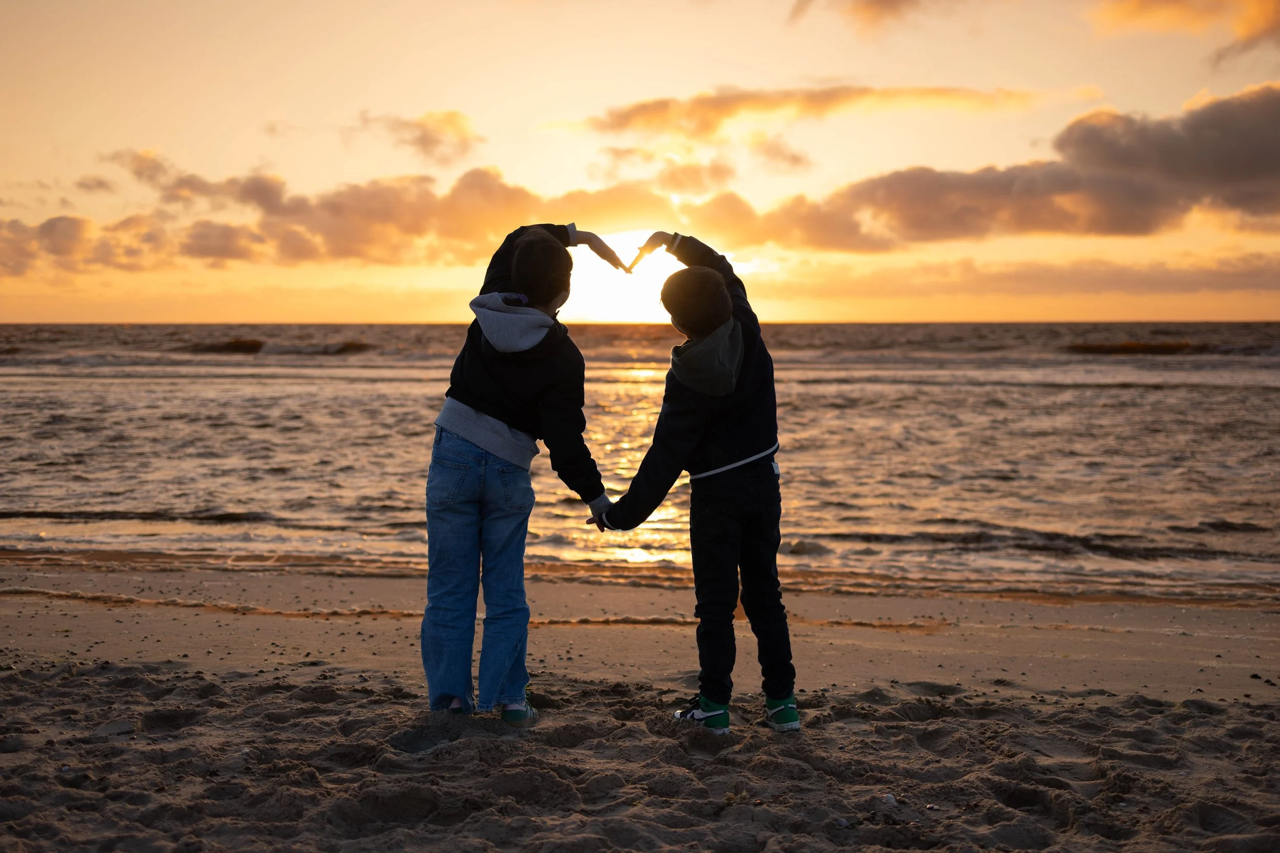 Zwei Kinder stehen am Strand, halten sich an den Händen und formen ein Herz mit den Armen vor Sonnenuntergang über dem Meer.