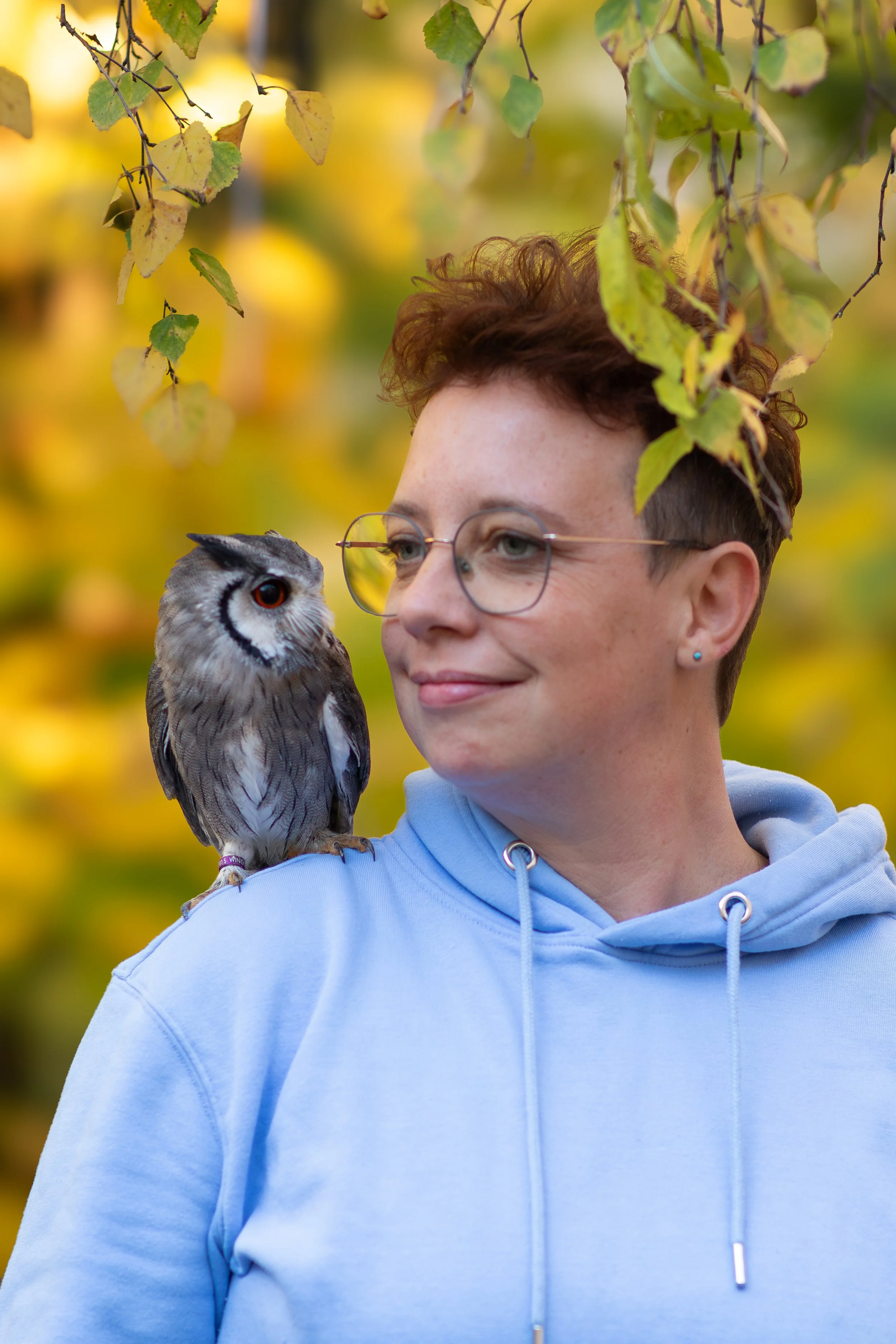 Junge Frau mit Kurzhaar, rötlich, und runden Brillen, trägt eine hellblaue Hoodie, steht im Freien mit grünen und gelben Herbstblättern um sie herum.