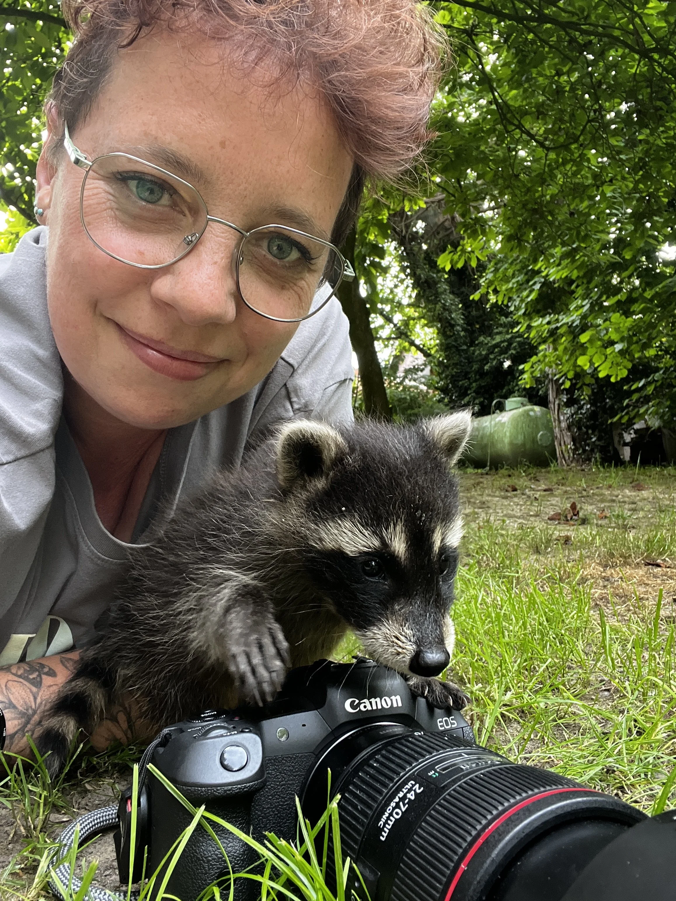 Eine Frau mit Brille und Tattoos, die im Garten auf dem Boden liegt, einen Hund hält und eine Kamera vor sich hat.