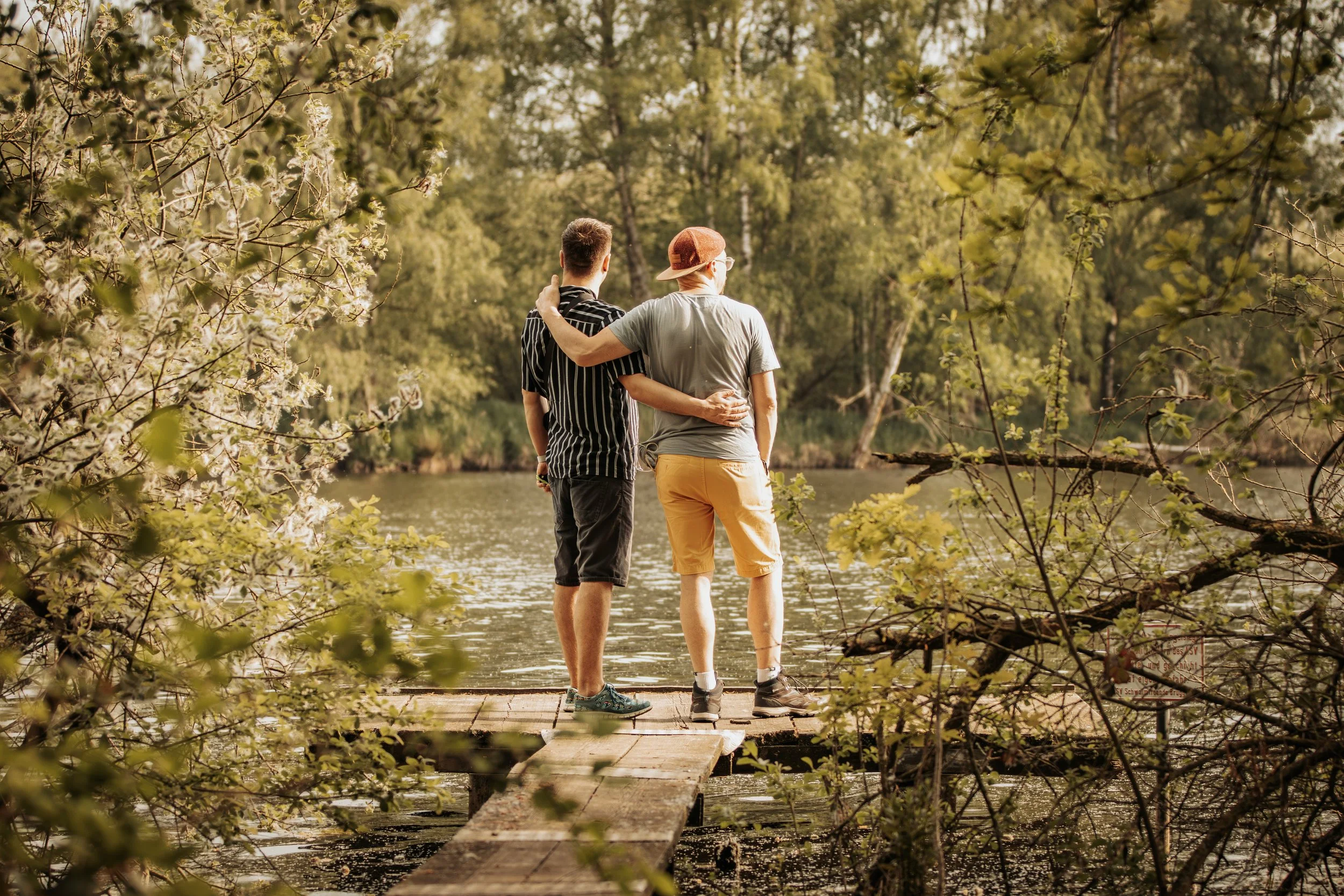 Zwei junge Männer stehen auf einem Holzsteg an einem Fluss im Wald. Sie umarmen sich und blicken auf die Wasseroberfläche, umgeben von grünen Bäumen.