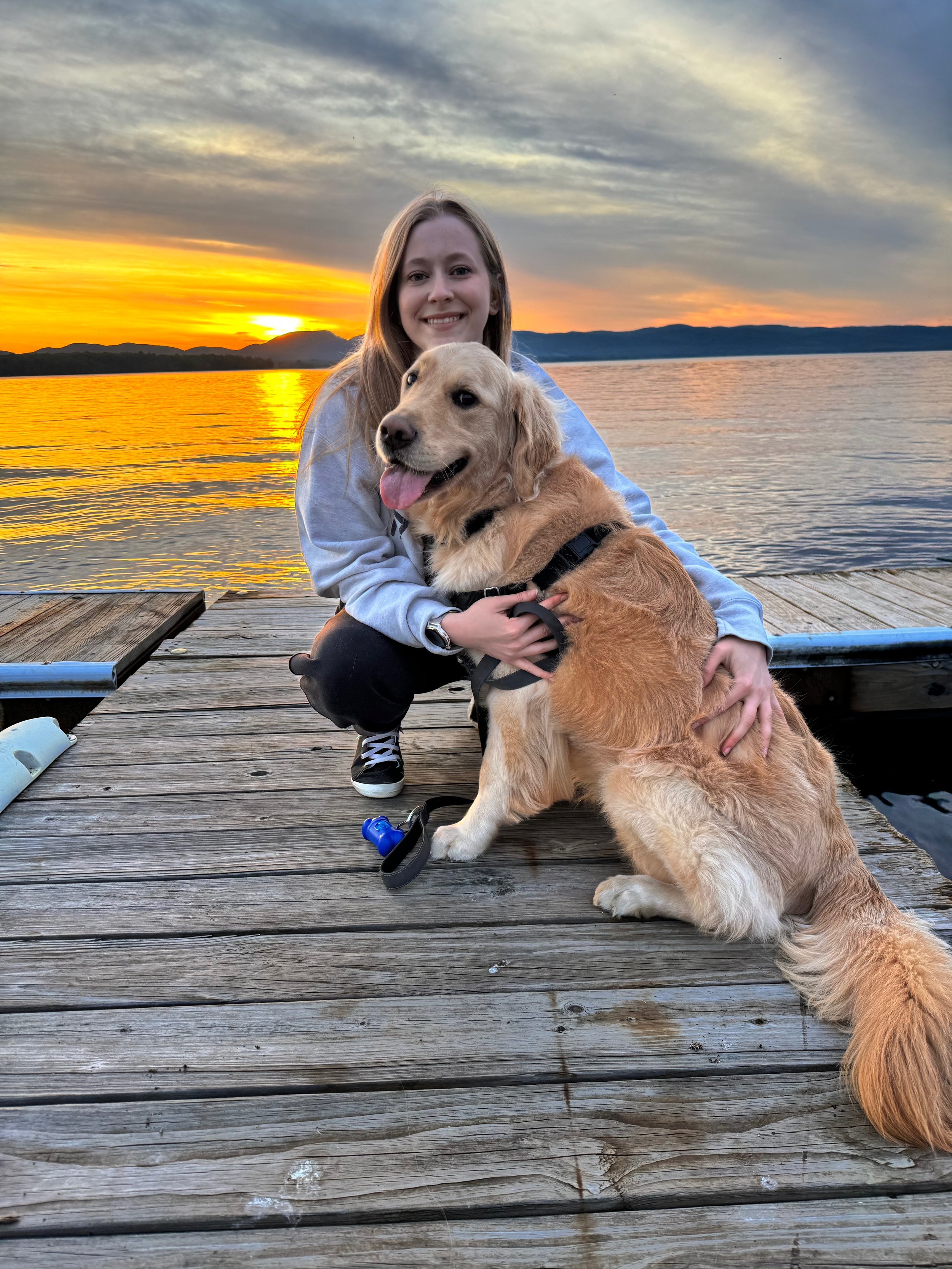 A young woman with long hair smiling and hugging a golden retriever puppy on a wooden dock by a lake at sunset.