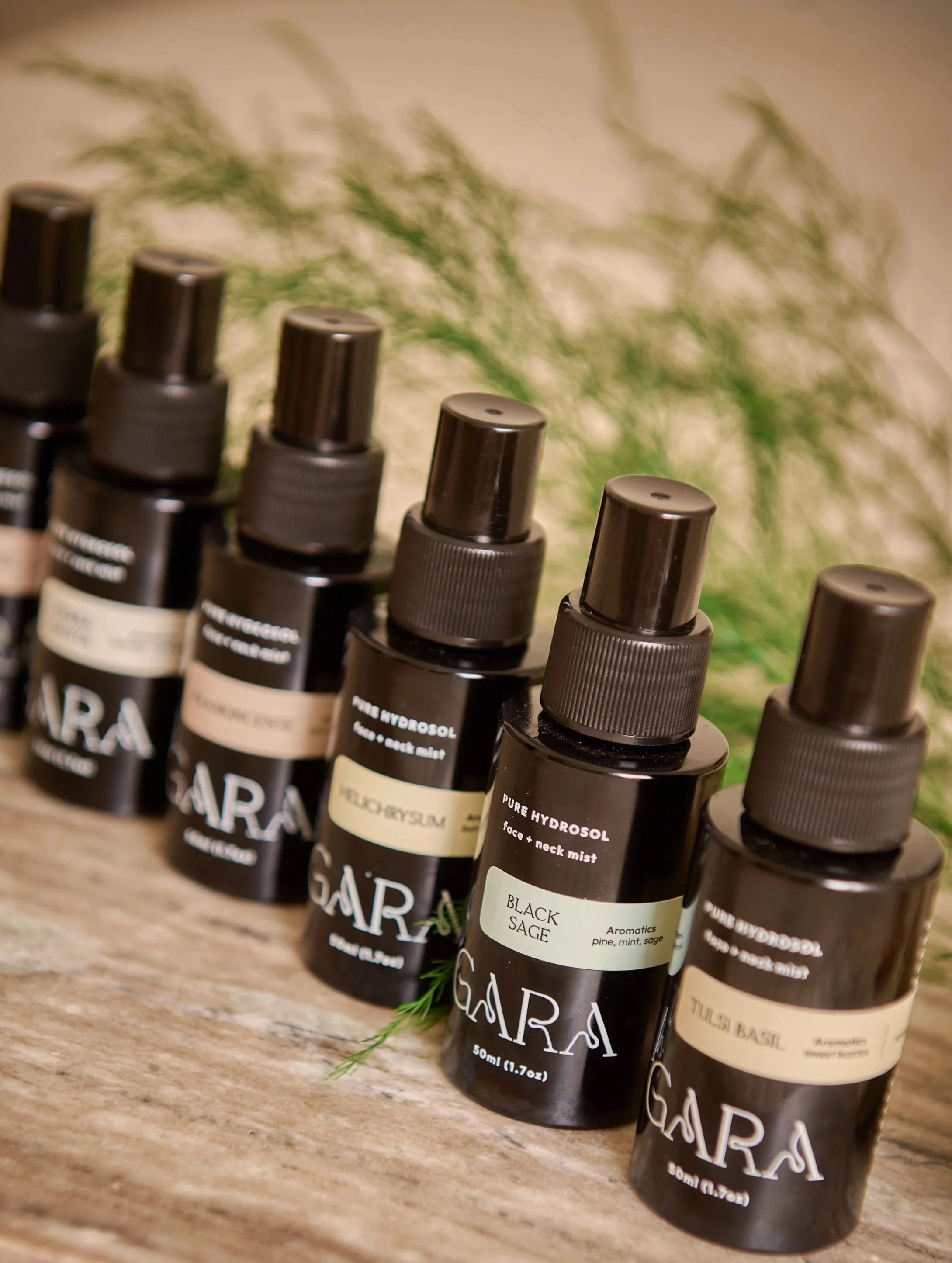 Multiple small dark bottles labeled with different scents including black sage, tulsi basil, and others, arranged on a wooden surface with green foliage in the background.