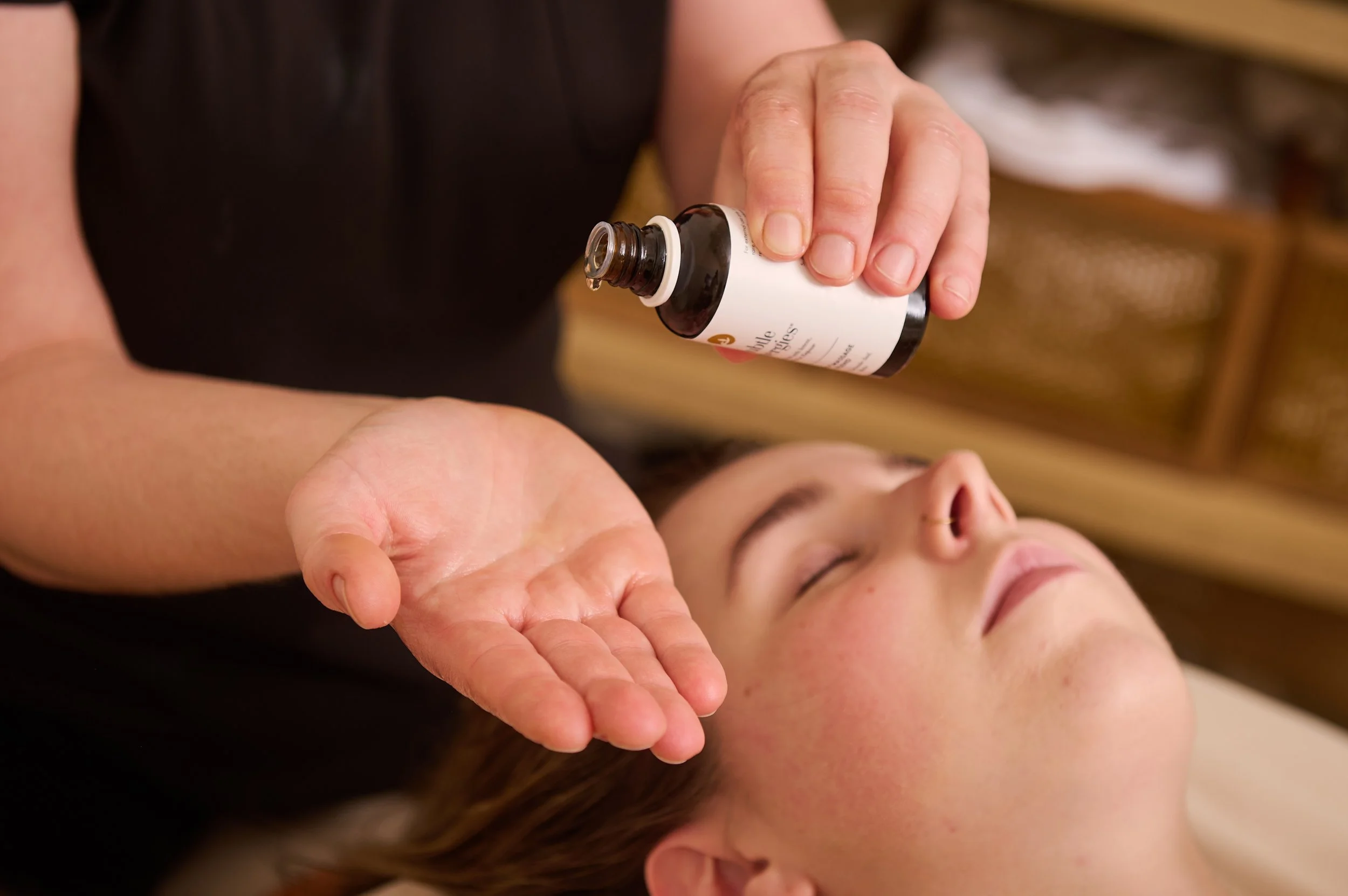A person holding a brown and white glass bottle with a white label over a person's forearm, who is lying down with closed eyes, possibly getting an eye treatment or massage.
