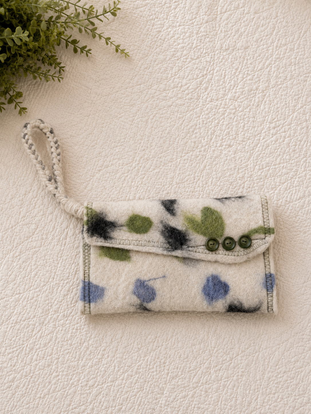 A felted wool pouch with a flap closure decorated with blue, green, black, and gray abstract shapes and three green buttons, lying on a textured white surface, with a green leafy plant in the upper left corner.