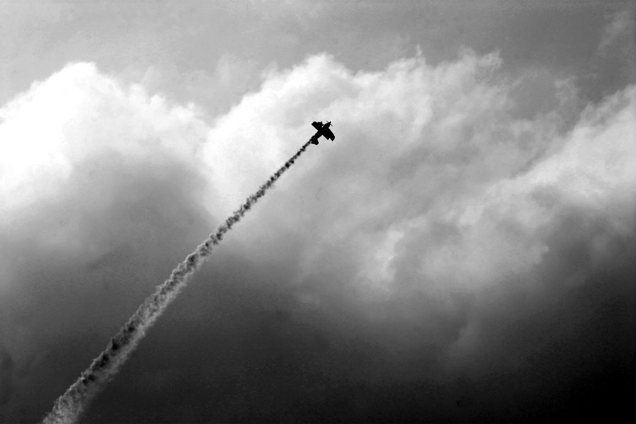 A silhouette of a small airplane flying in the sky, leaving a trail of smoke or vapor behind it, with clouds in the background.