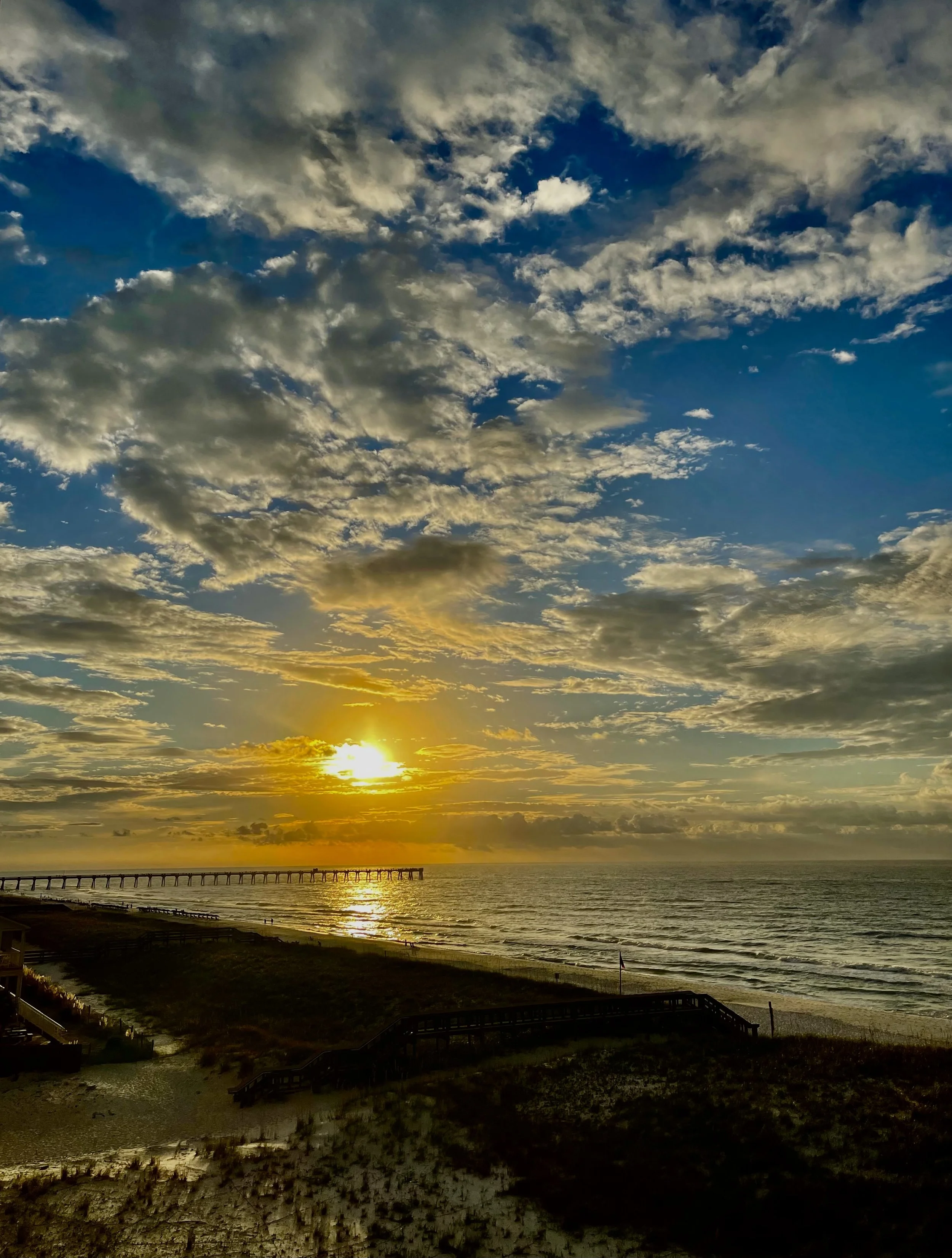 Sunset over a cloudy sky with a pier extending into the ocean and a sandy beach in the foreground.