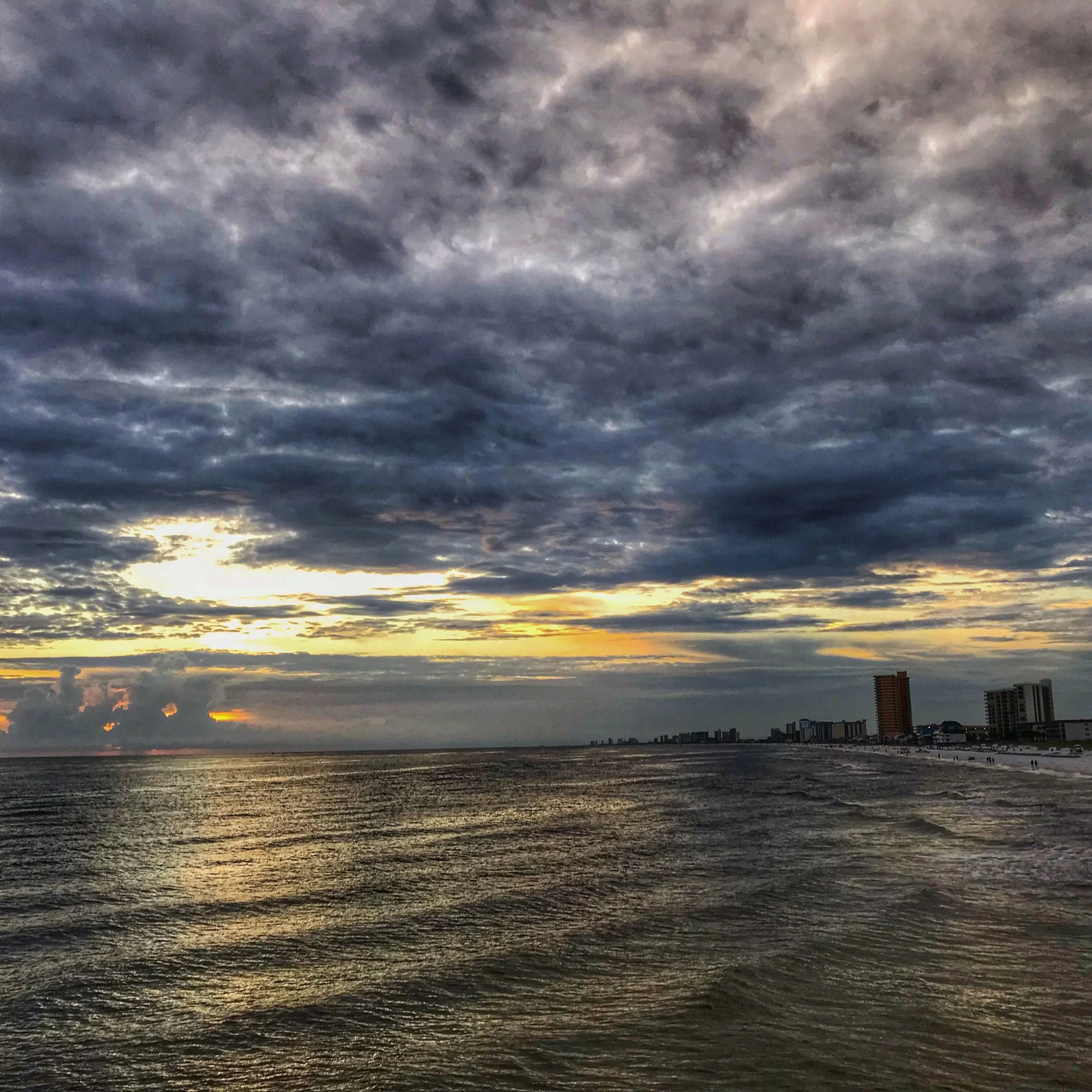 Sunset over the ocean with dark clouds in the sky and city buildings in the distance.