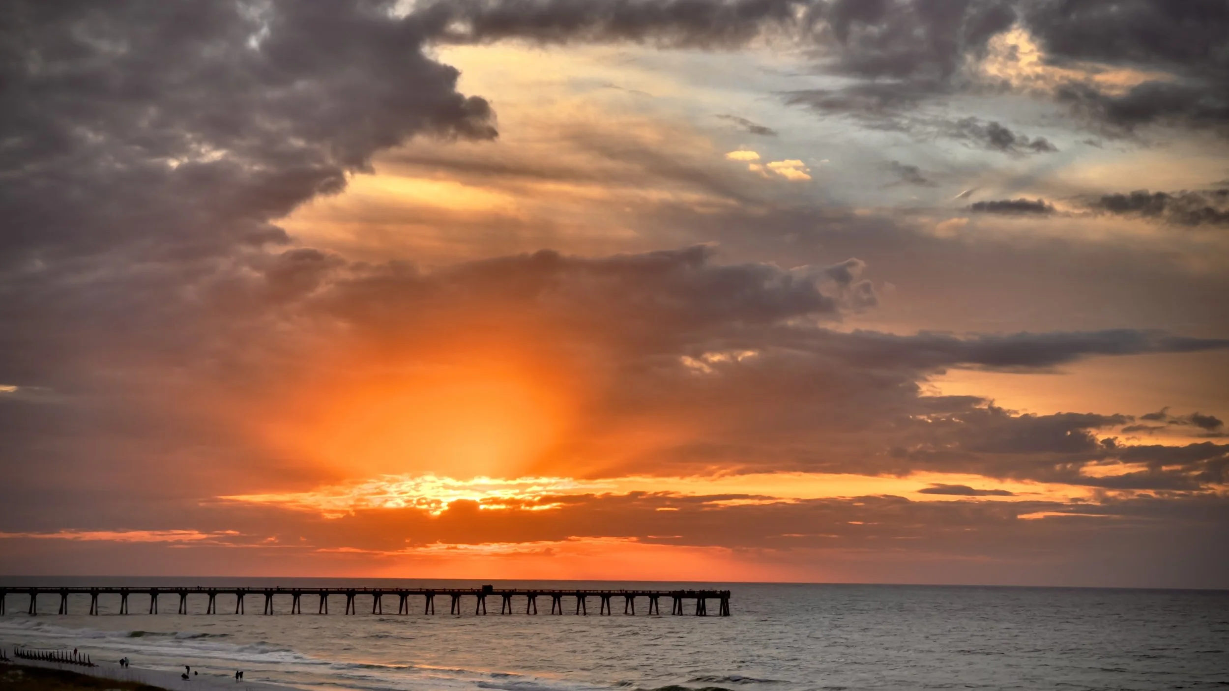 Sunset over the ocean with a dramatic sky of dark clouds and orange light, a pier stretching into the water, and a sandy beach with a few people.