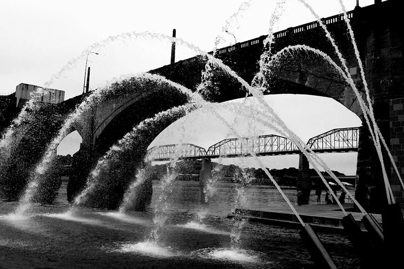 Black and white photo of a bridge over a river with water fountains spraying through arches.