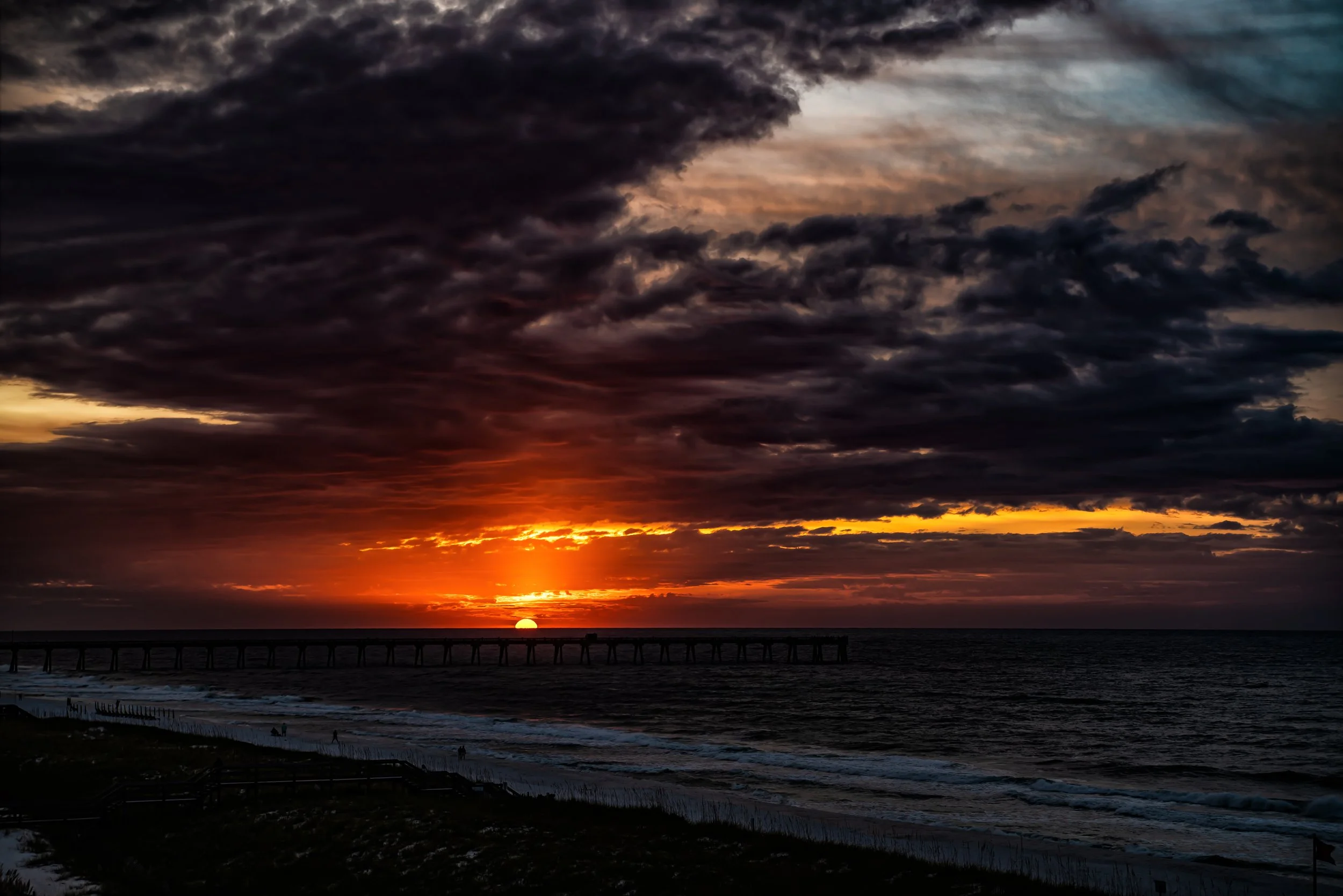 Sunset over the ocean with dark, layered clouds and a pier extending into the water.