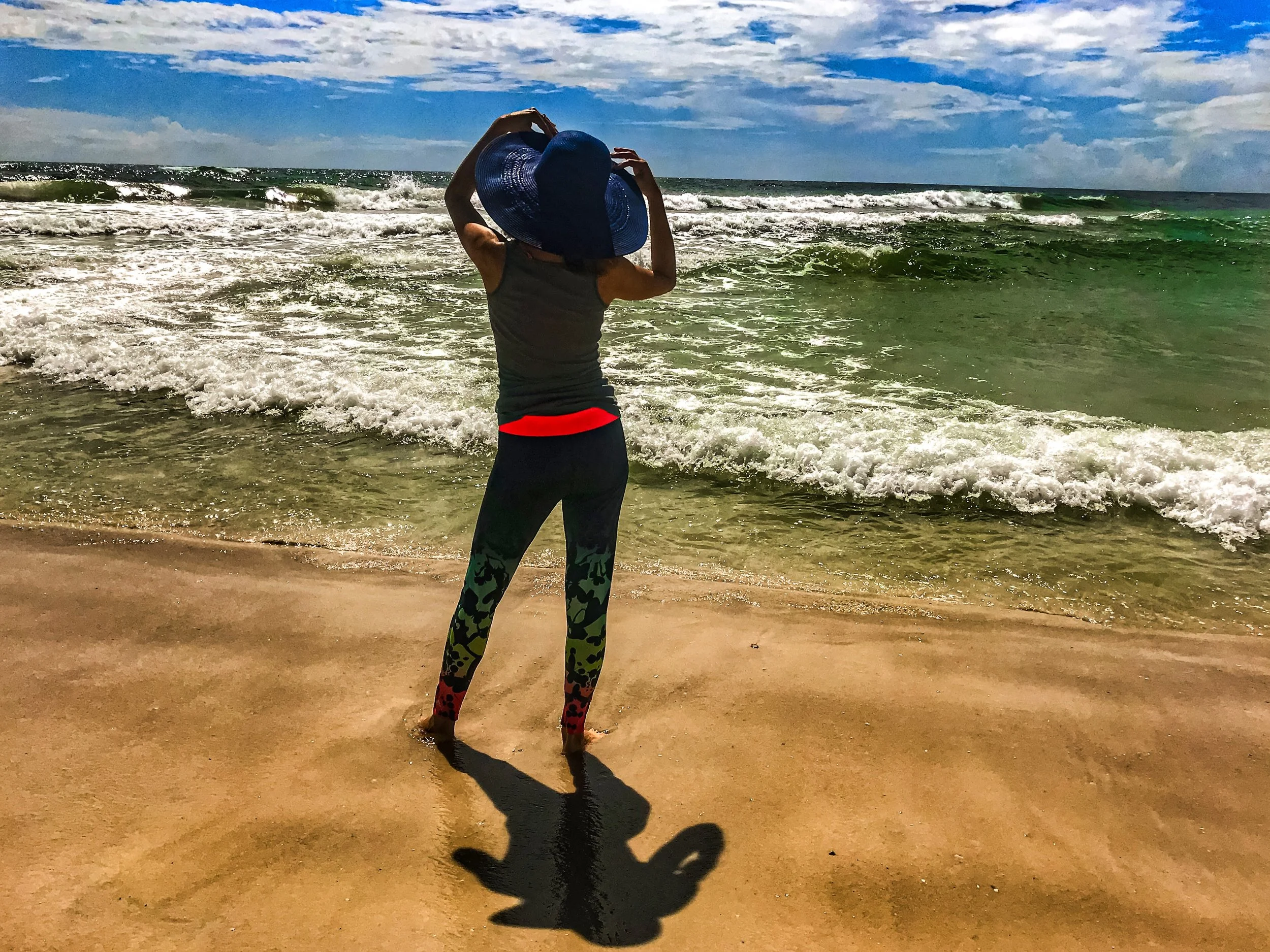 Person standing on sandy beach with waves and blue sky, wearing a large blue sun hat, gray tank top, and colorful leggings, casting a shadow.