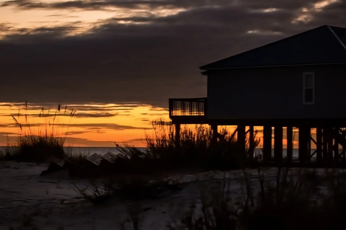 A house on stilts at the beach during sunset with clouds and orange hues in the sky.