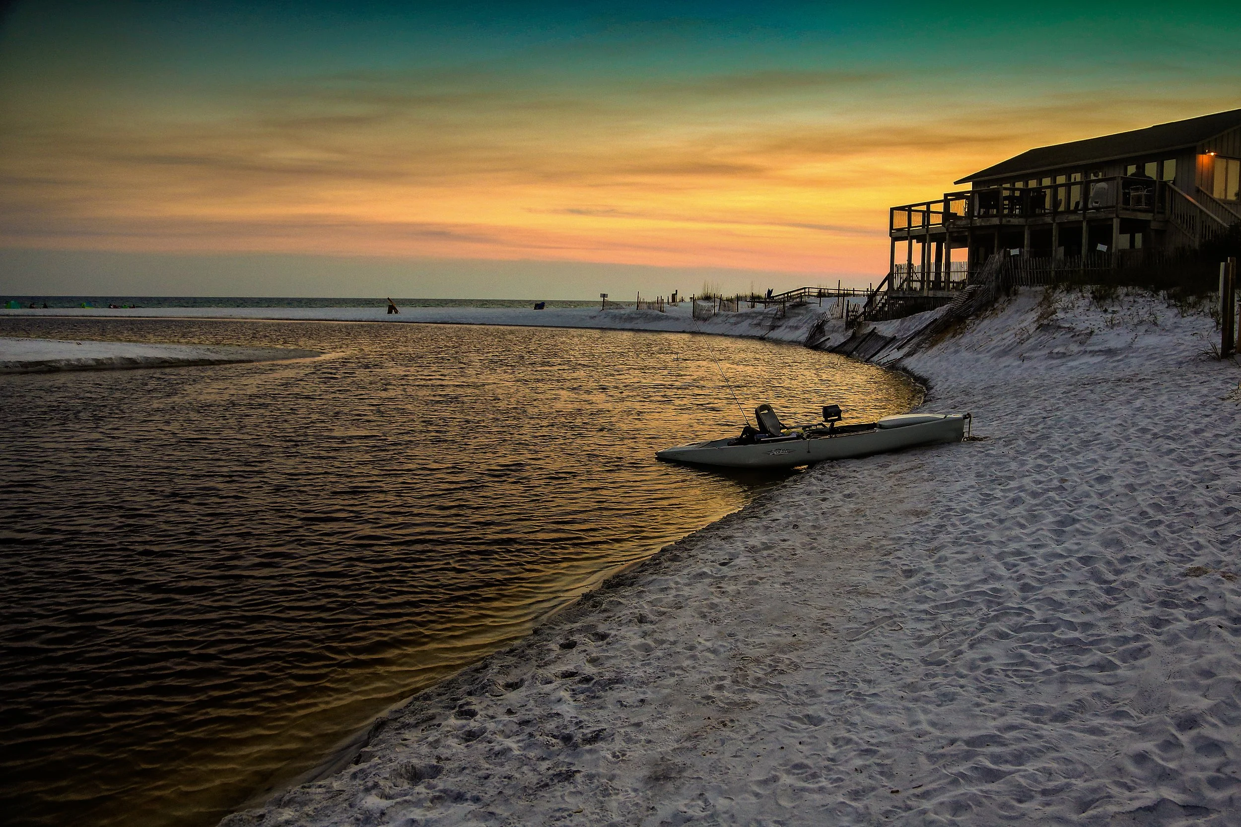 Sunset over a beach with a house on stilts, a small boat on the sand, and a calm ocean with a few people in the distance.