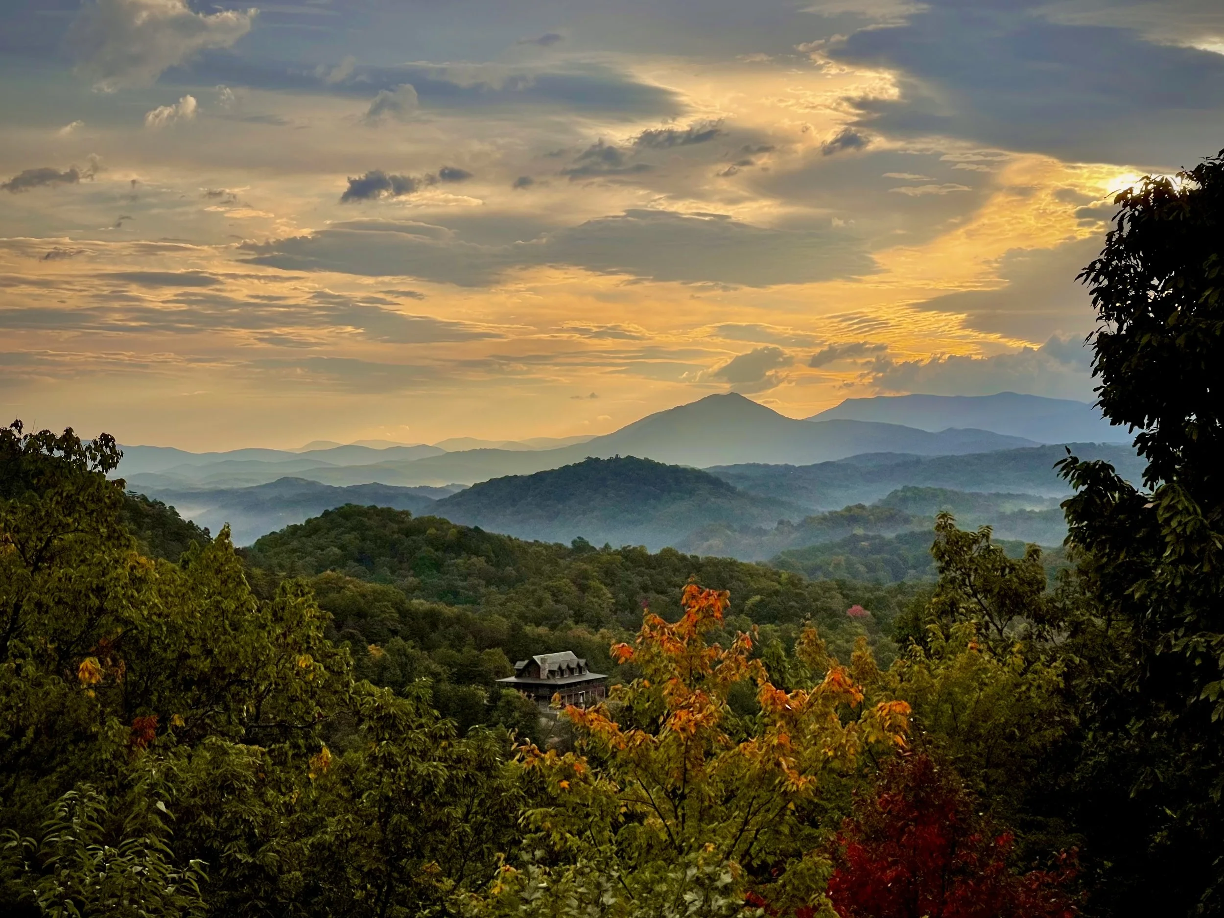 A scenic view of a sunset over a mountainous landscape with a house in the valley, framed by green and orange trees in the foreground.