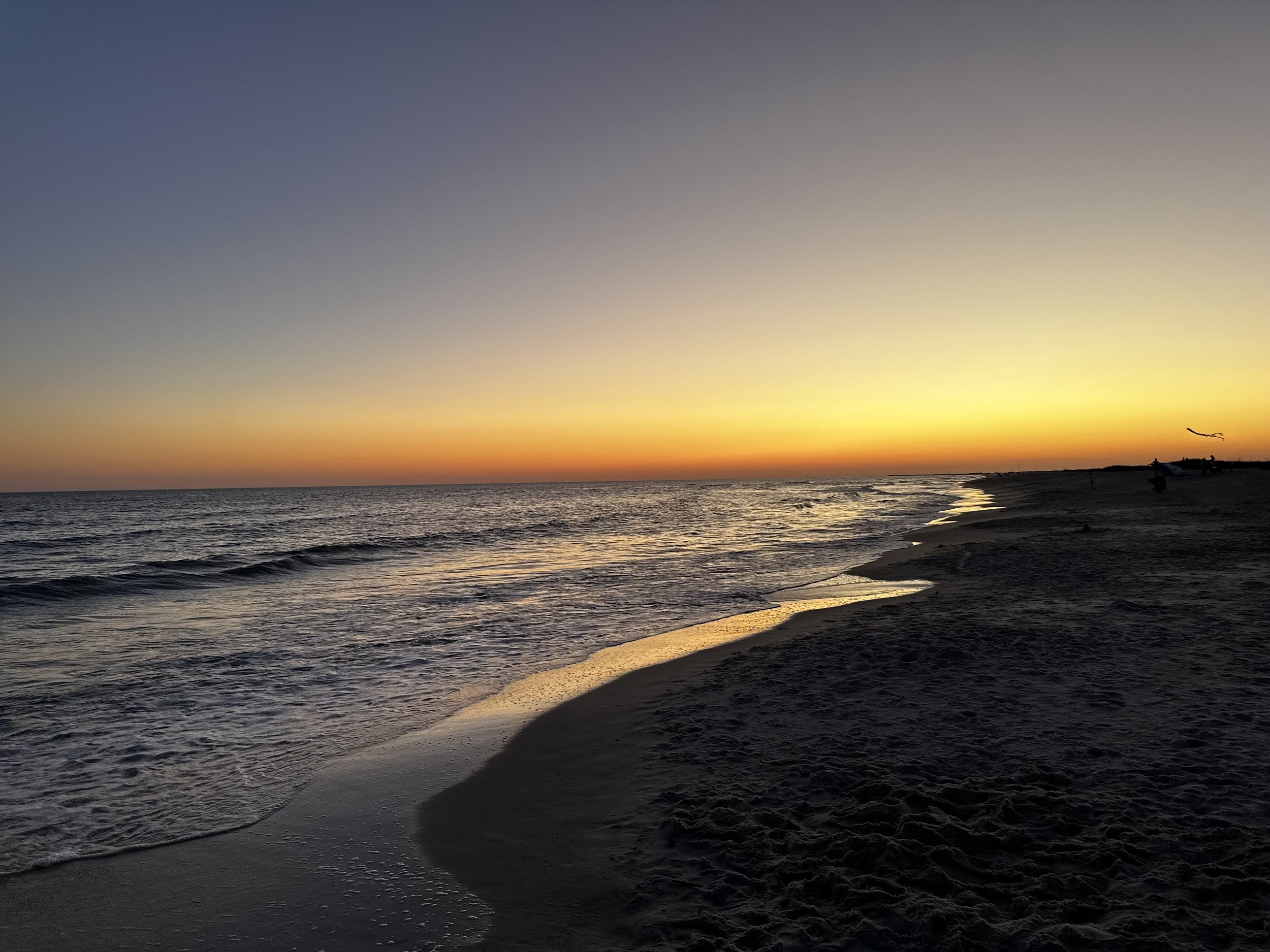 A sunset over the ocean with a few people and a kite in the distance on the beach.
