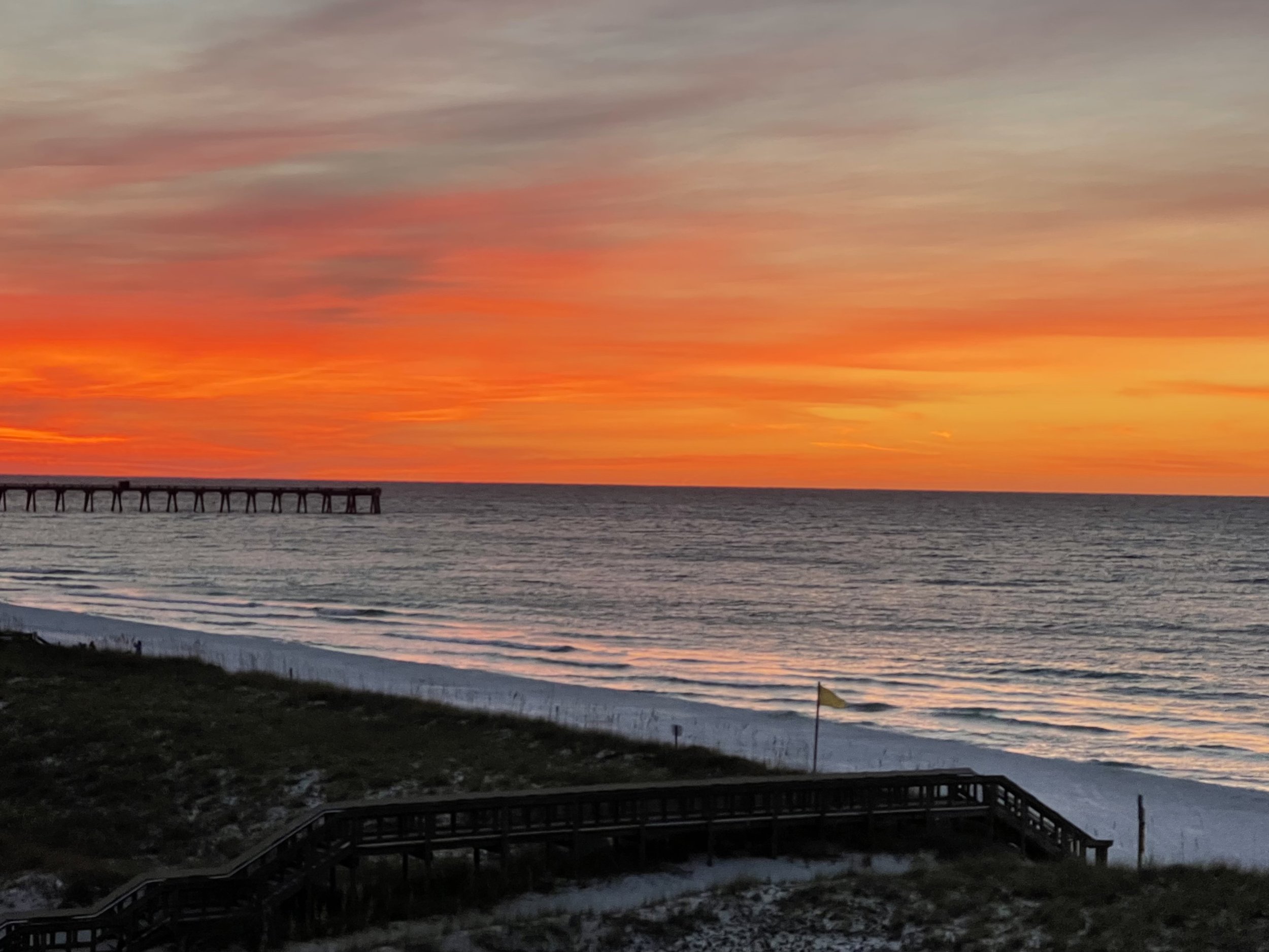 Sunset over the ocean with a colorful sky of orange, pink, and purple hues. A pier extends into the water on the left side of the image, and a small flag is visible near the bottom right.