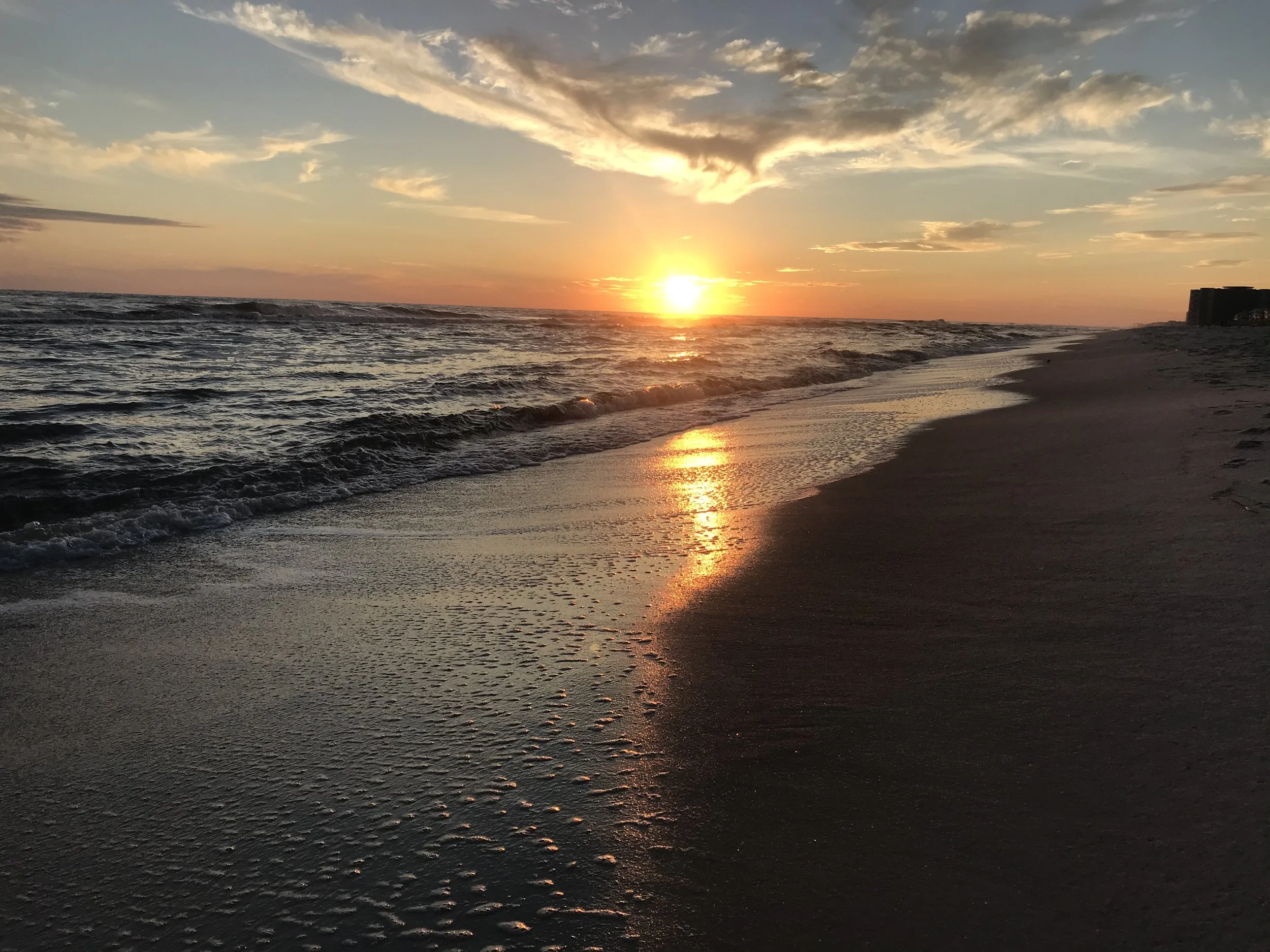 A sunset over the ocean with the sun near the horizon, reflecting orange and yellow hues onto the water and sky, with clouds scattered above and a sandy beach in the foreground.