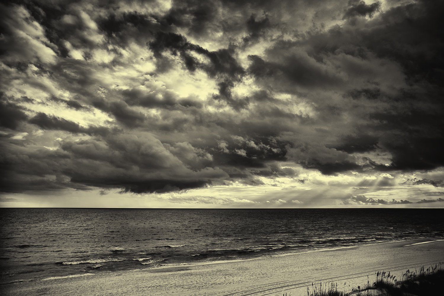 Black and white photograph of a beach with ocean waves and dark storm clouds overhead.