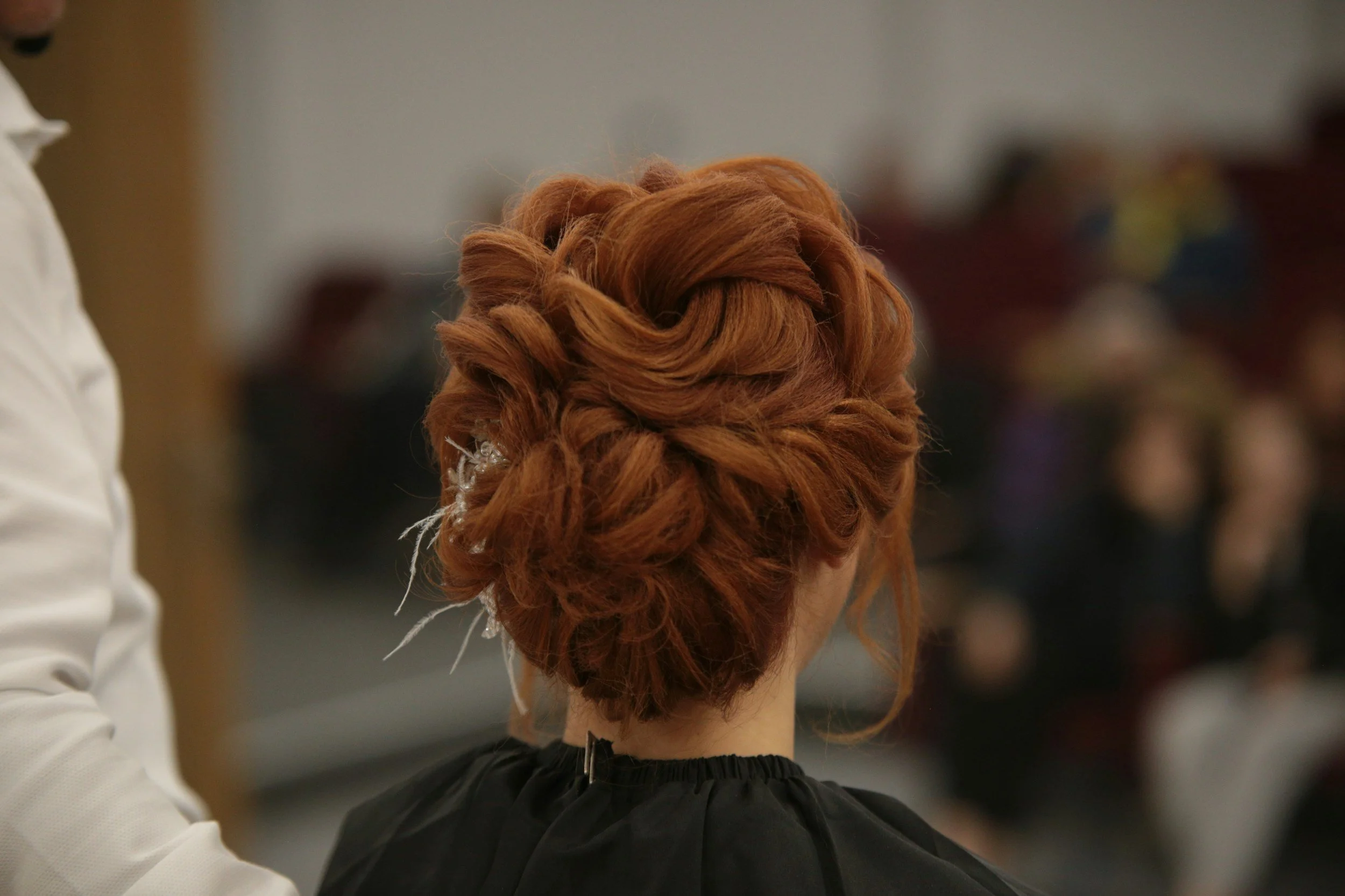 The back of a woman with styled red hair, possibly at a hair event or salon, with blurred background.