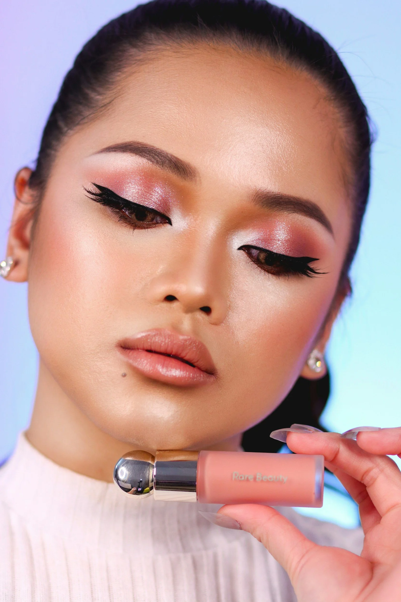 Close-up of a woman with well-groomed eyebrows and eye makeup, holding a bottle of Rare Beauty liquid blush near her face.