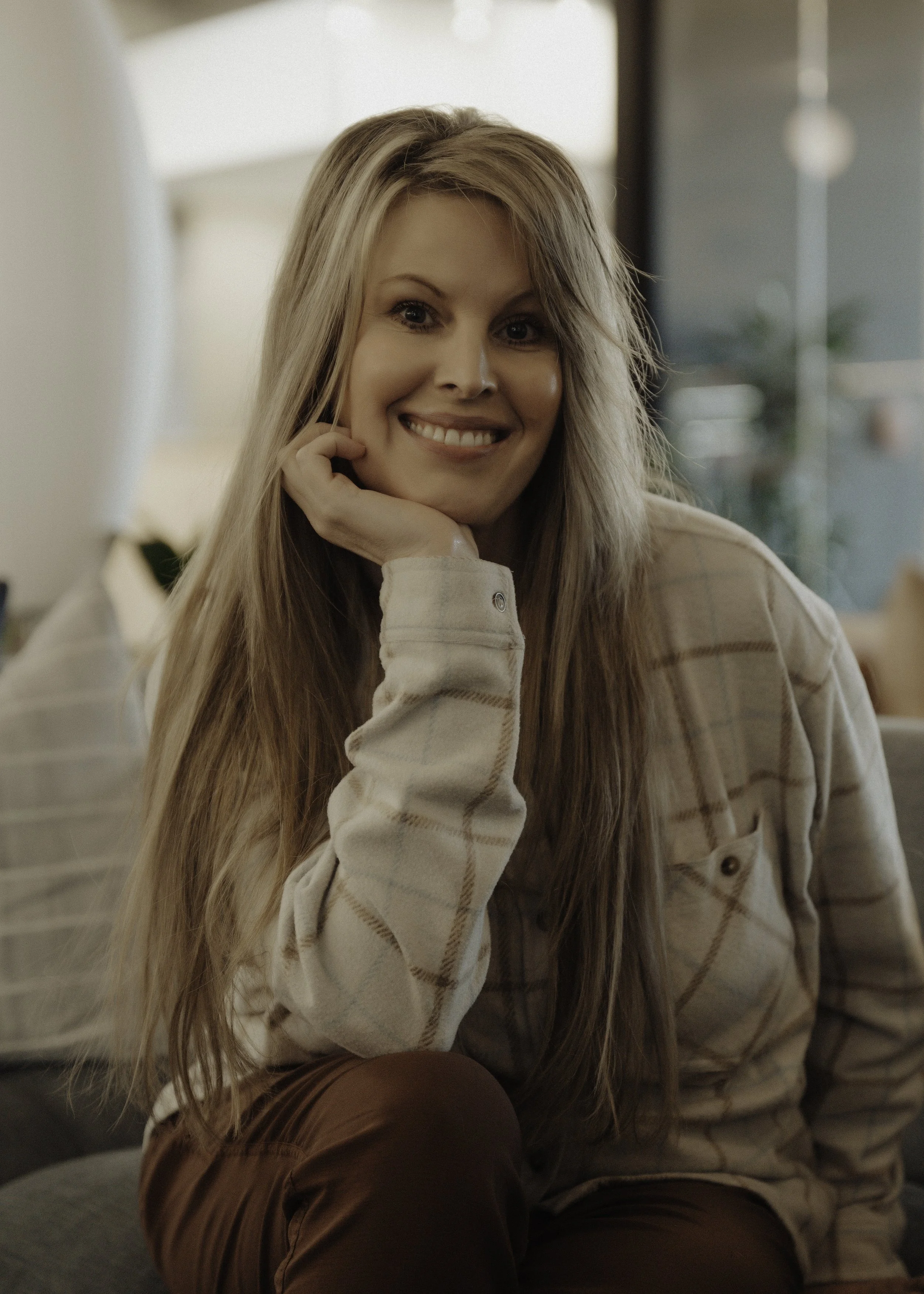 A young woman with long blonde hair, smiling and resting her chin on her hand, sitting indoors with a blurred background.