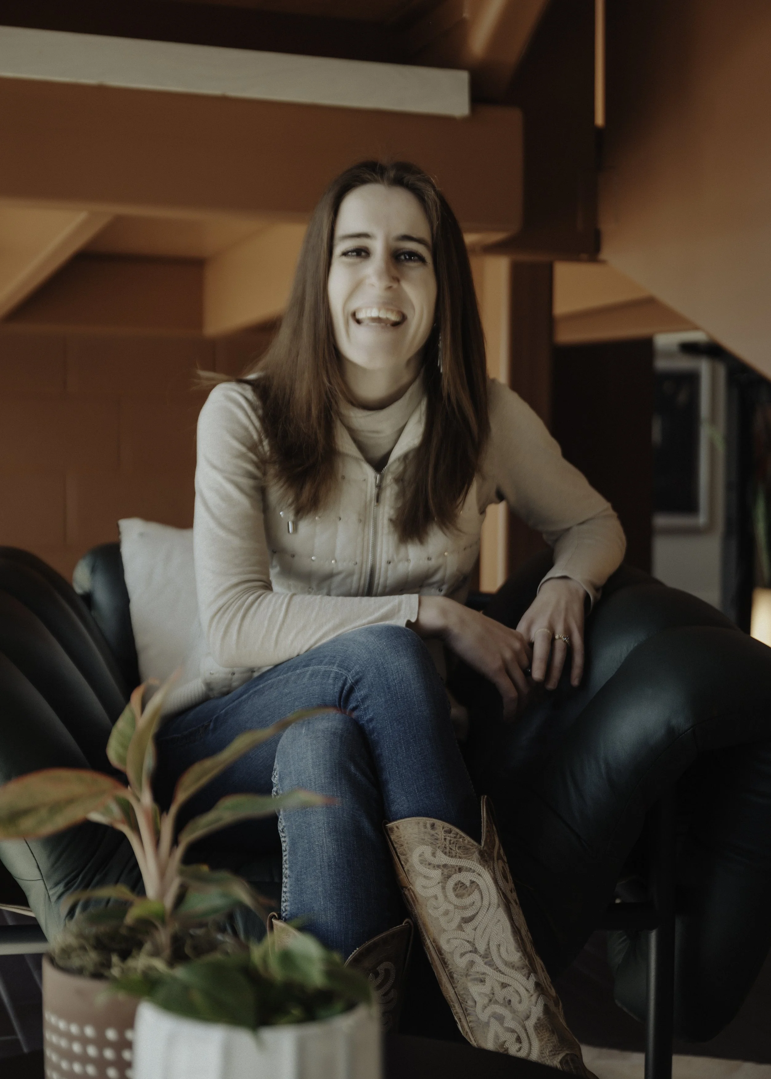 A woman with long brown hair smiling and sticking her tongue out, sitting on a black leather couch in a warmly lit room with brown walls. She is wearing a beige zip-up jacket and blue jeans, with a decorative brown and beige patterned bag on her lap. In the foreground, a potted plant with green leaves is partially visible.