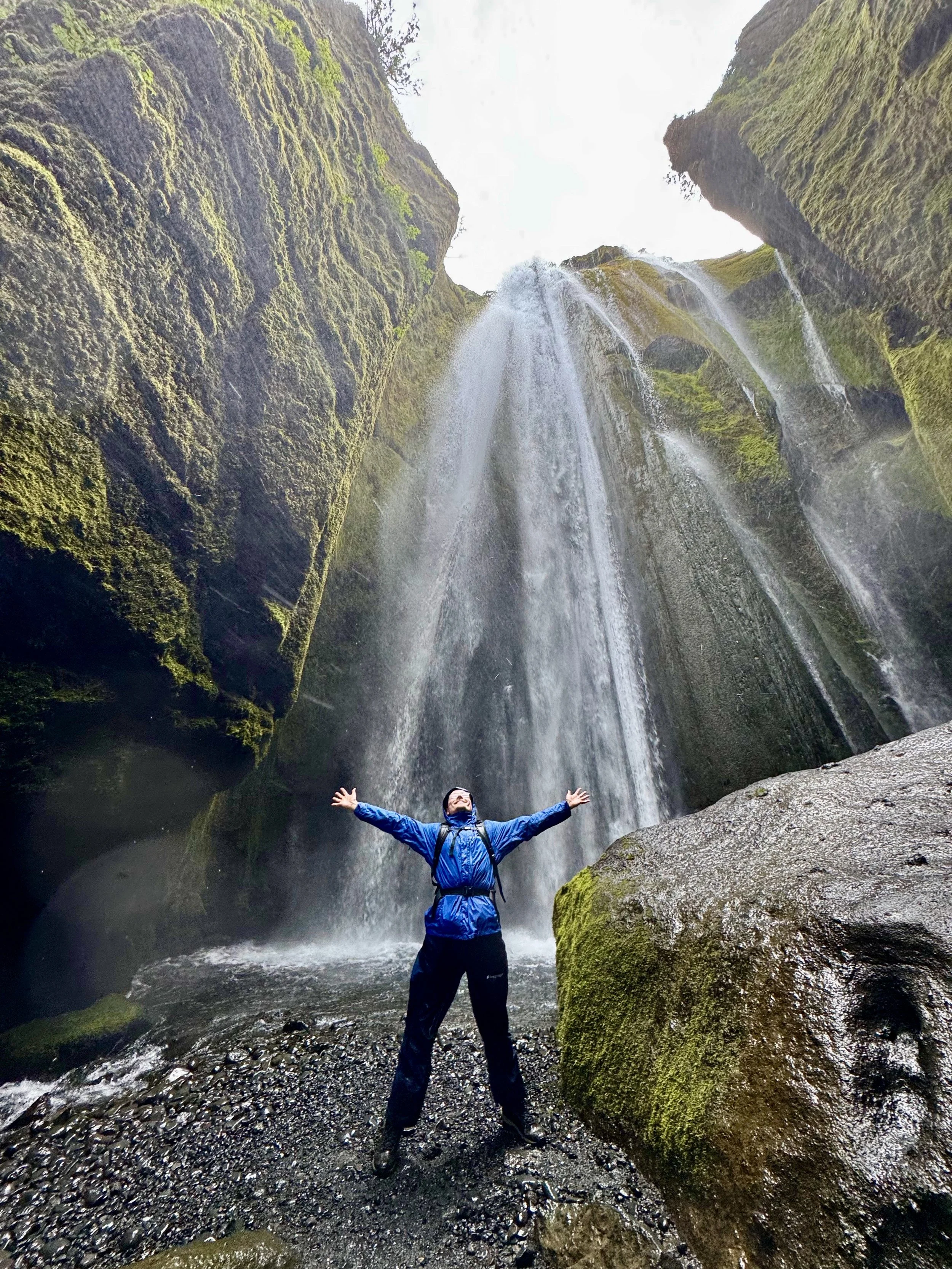 Person standing with arms outstretched in front of a waterfall inside a narrow canyon with moss-covered rocks and water flowing down from above.
