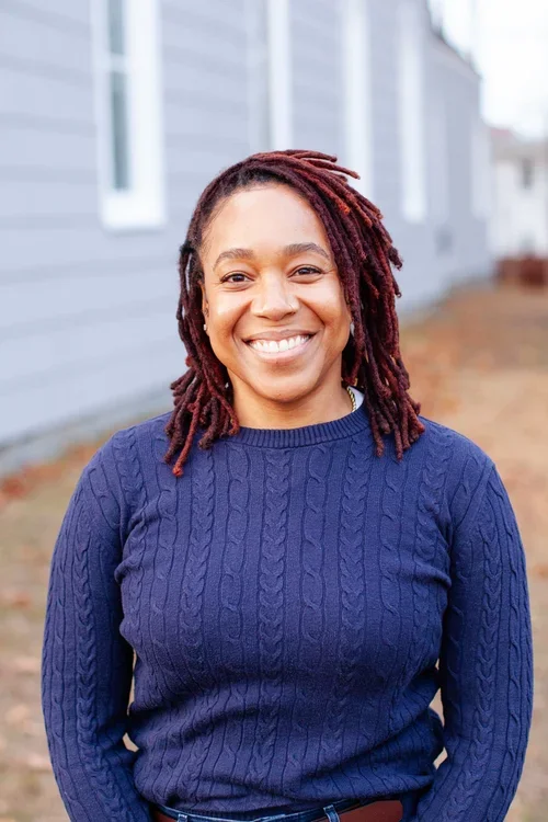 A woman with red dreadlocks smiling outdoors in front of a gray house, wearing a blue cable-knit sweater.
