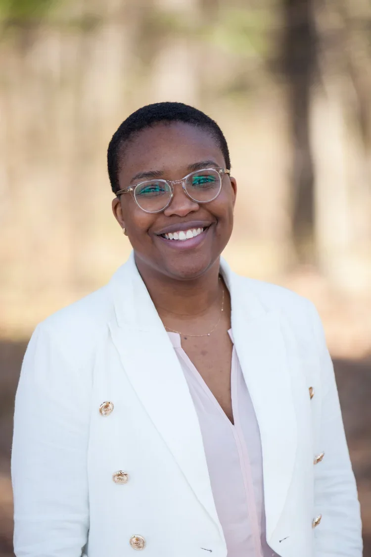 A smiling woman with short black hair, glasses, and a white blazer standing outdoors with blurred trees in the background.