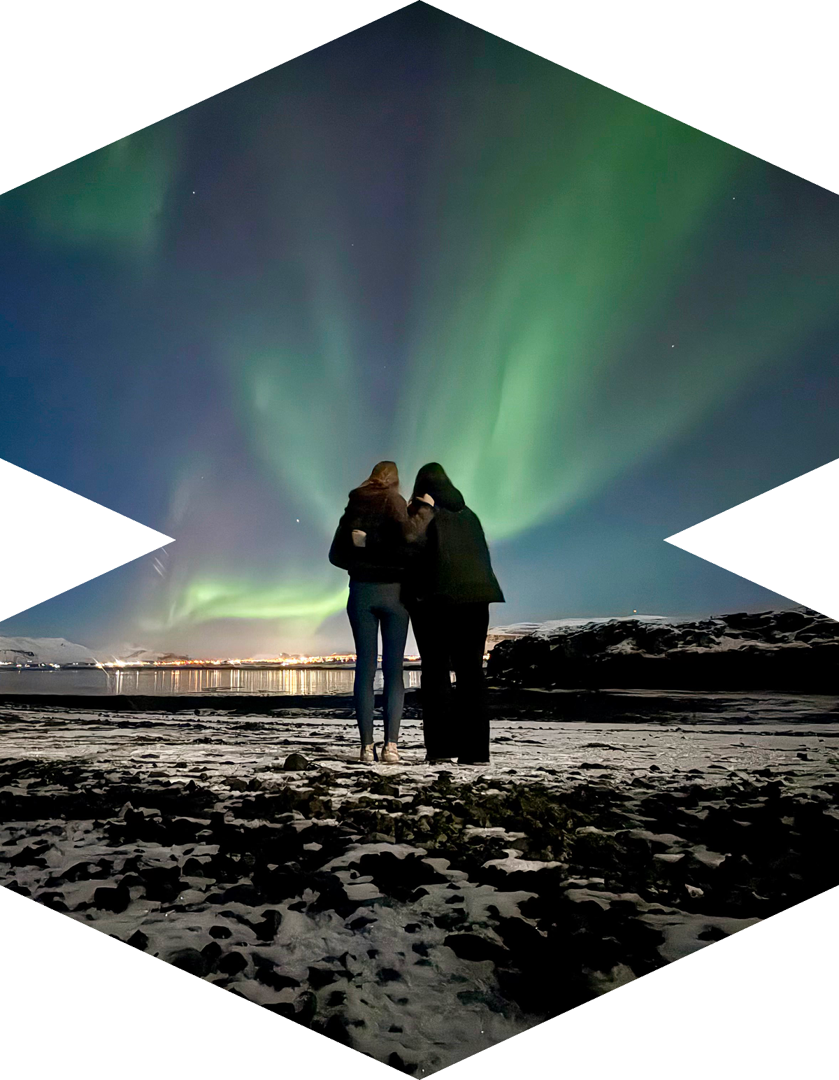 Two people stand close together on a snowy landscape, watching the Northern Lights display vivid green and blue auroras in the night sky above a body of water.