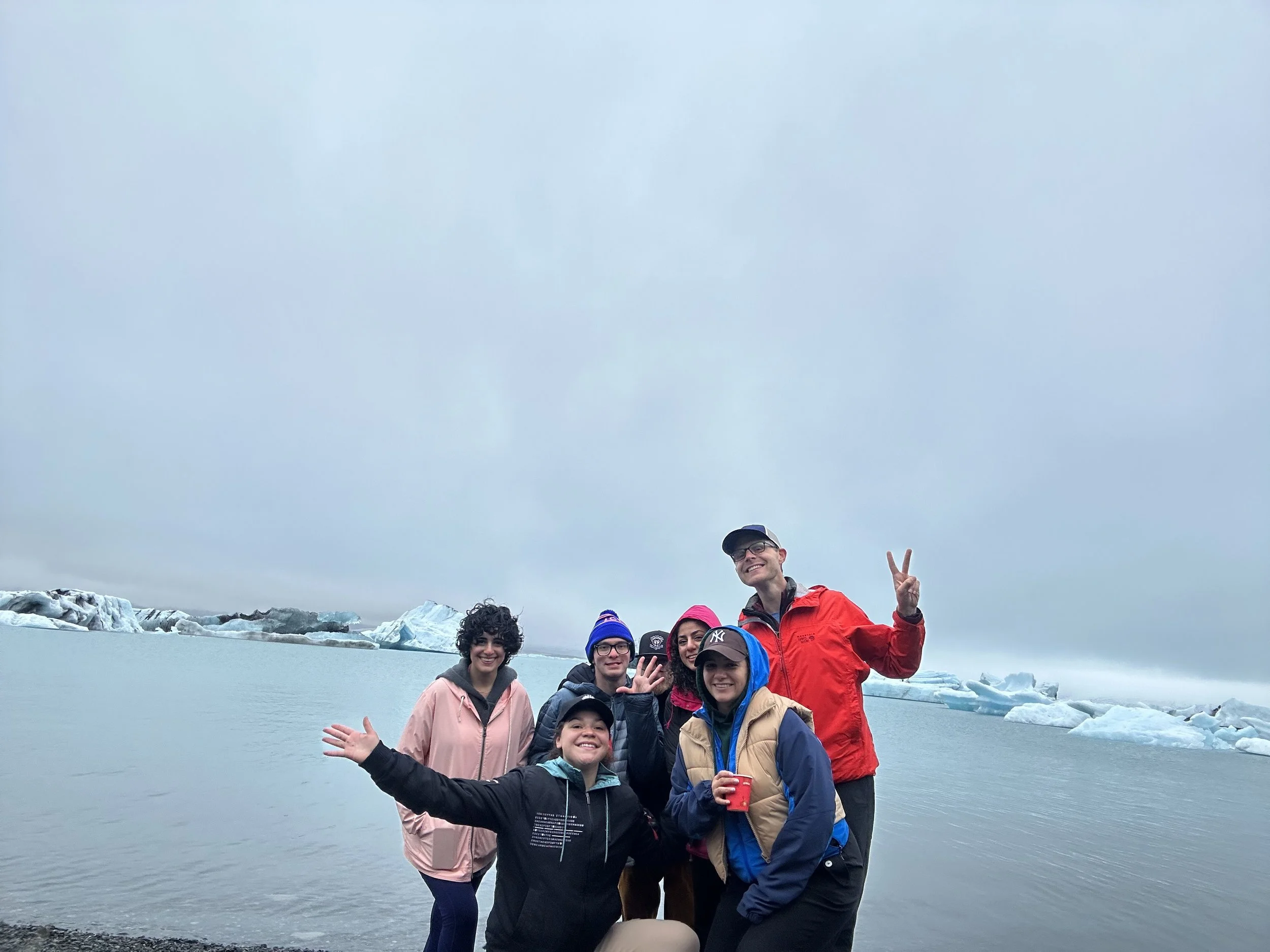 A group of people standing on a rocky shoreline with ice and a glacier in the background, under a cloudy sky. They are smiling and posing for the photo, some making peace signs.
