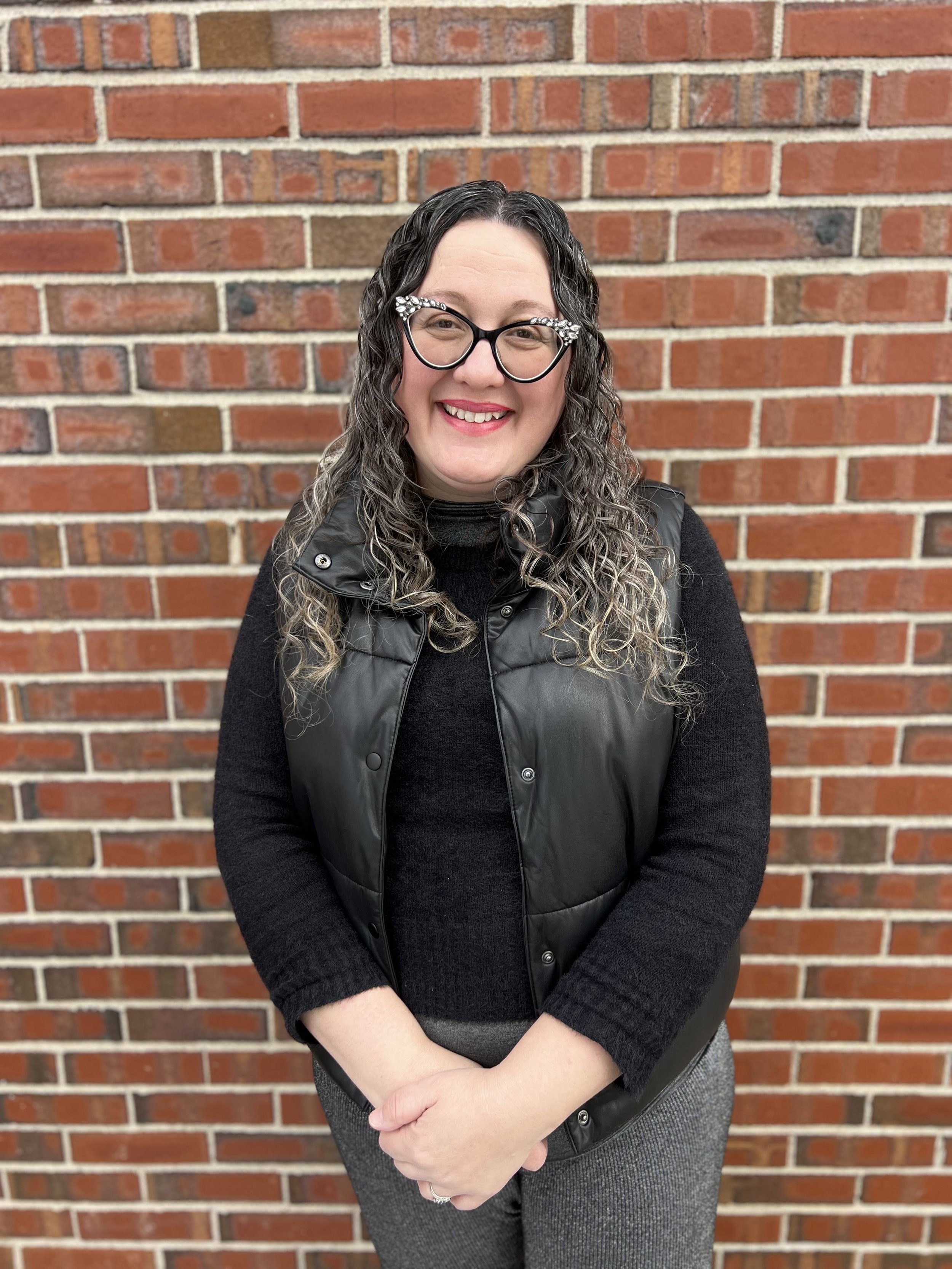 A woman standing in front of a brick wall, smiling, wearing glasses with floral embellishments, a black top, and a black vest.