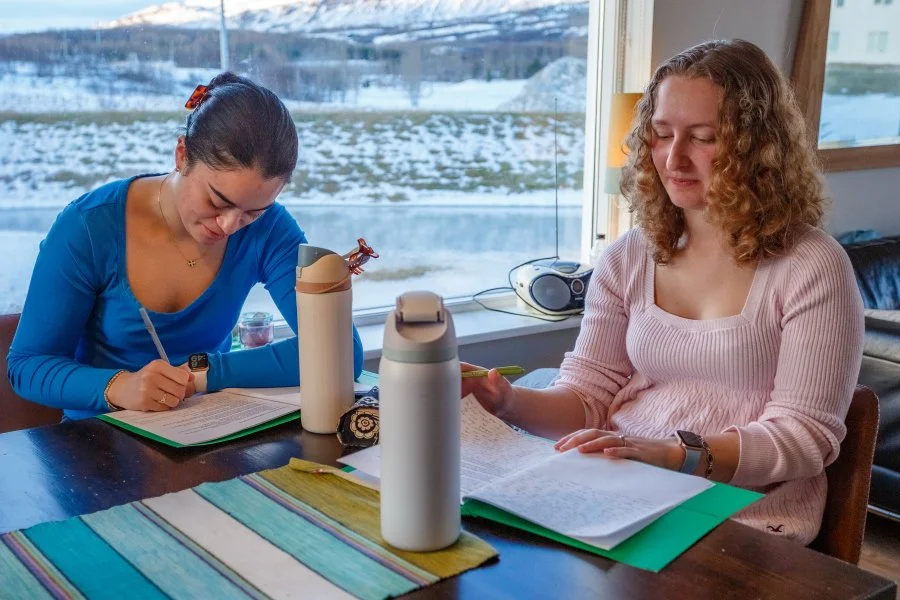 Two women are studying or working at a wooden table by a large window with snow outside. One woman in a blue top is writing, and the other in a pink top is reading or reviewing papers. Water bottles and a small speaker are on the table.