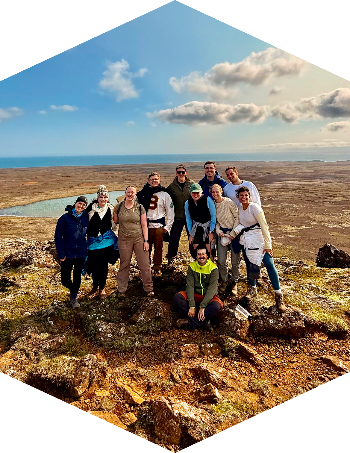 Group of eleven young adults standing on a rocky hilltop with a scenic landscape including water, open plains, hills, and a partly cloudy sky in the background.
