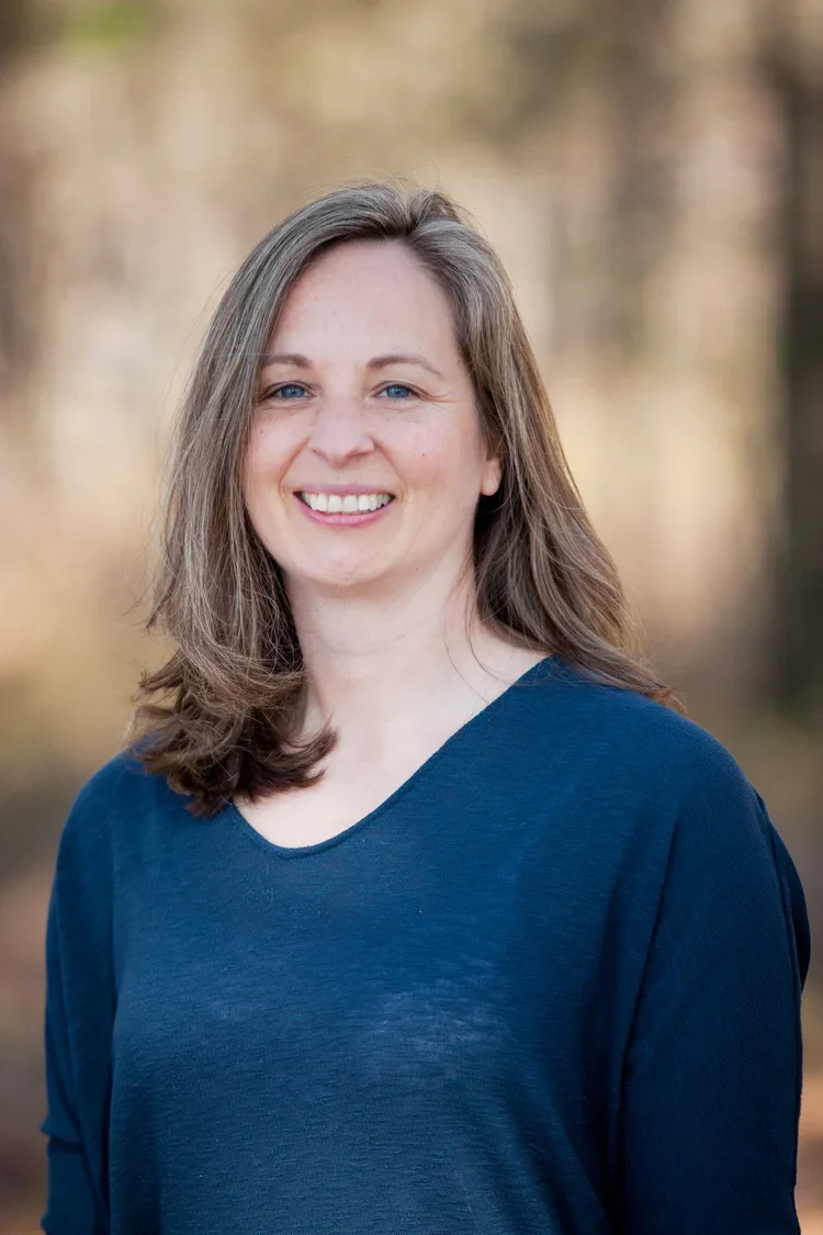 A woman with shoulder-length brown hair and blue eyes smiling, wearing a navy blue top, outdoors with blurred trees in the background.
