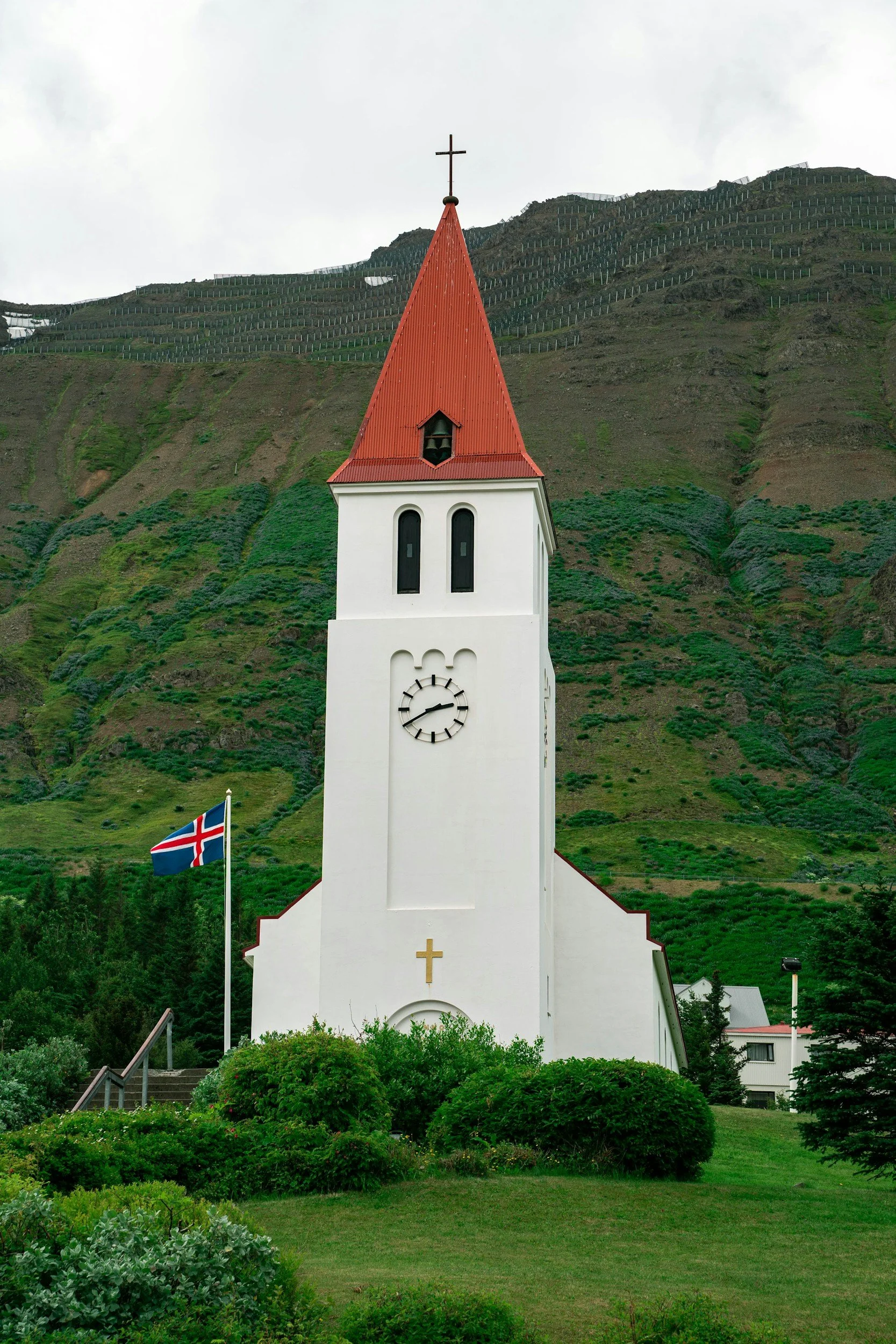 White church with red steeple and cross, situated at the base of green, hilly landscape with vineyards, Icelandic flag in lawn, and overcast sky.