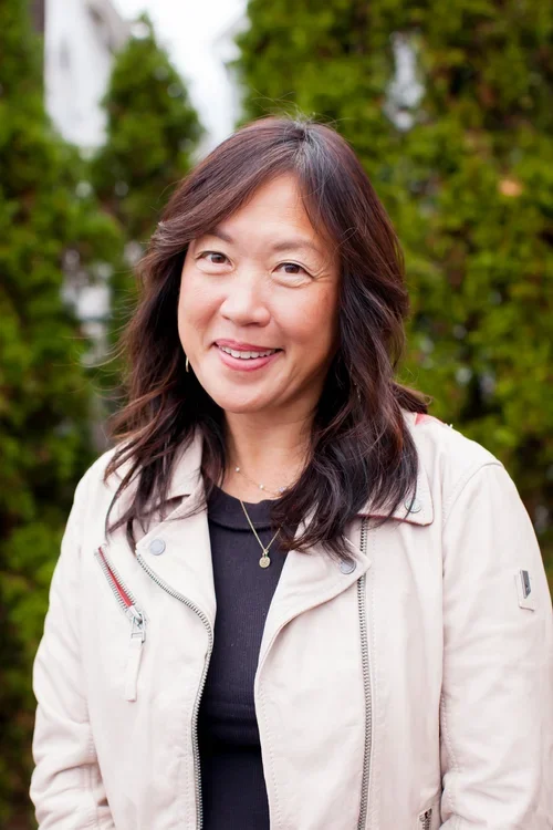 smiling woman with dark hair outdoors, wearing a light-colored jacket and black top, with green trees in the background