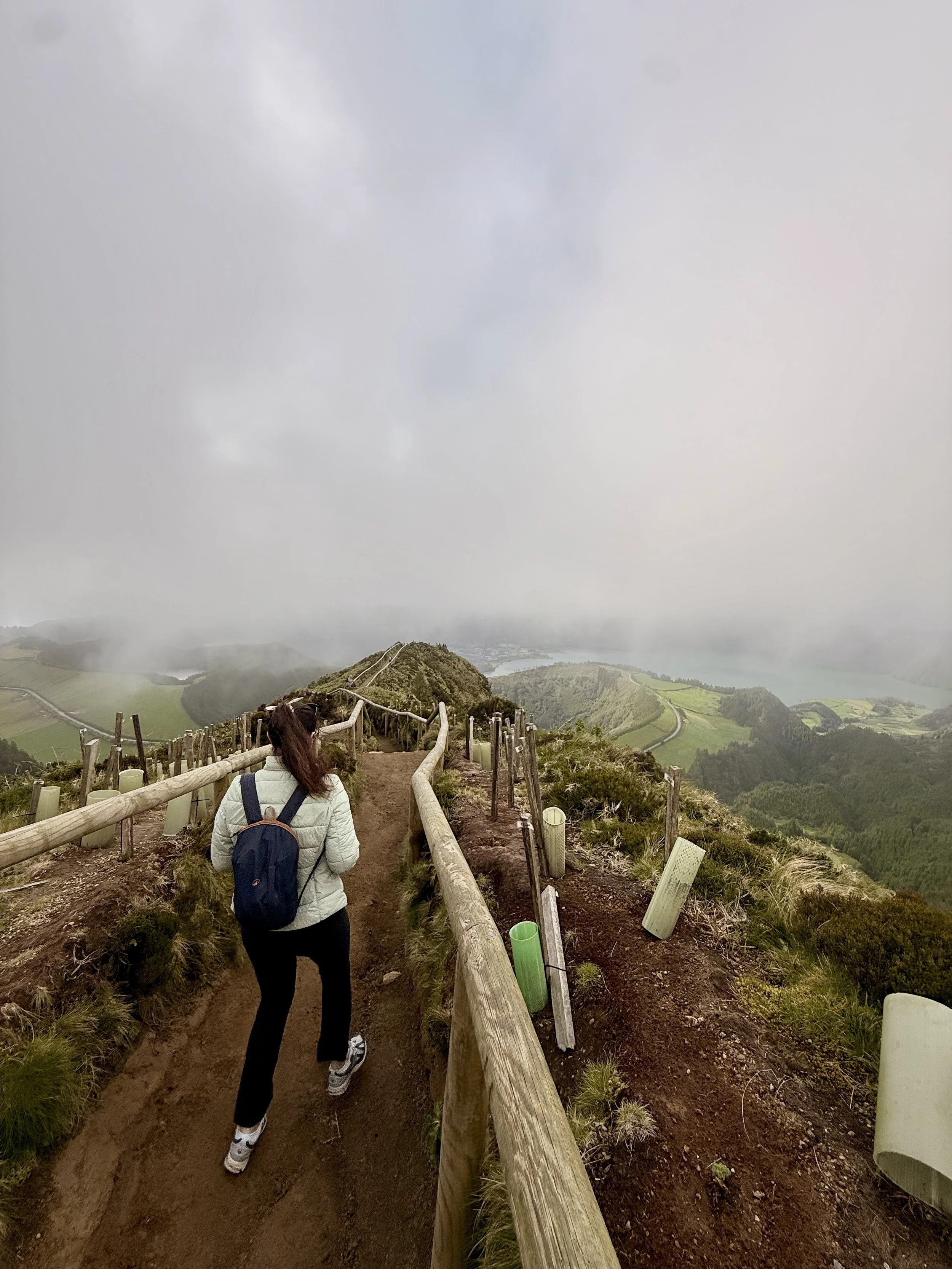 A person with a backpack walking down a dirt trail on a mountain in the Azores, with a view of foggy, green hills and valleys in the background.