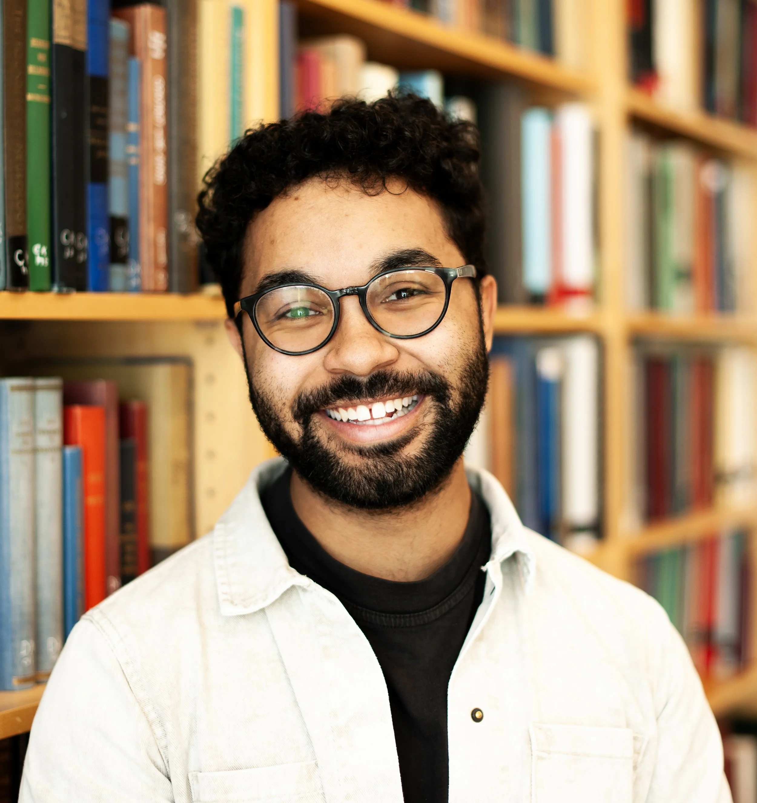 Close-up of a smiling man with glasses, curly hair, and beard, standing in front of a bookshelf filled with colorful books.