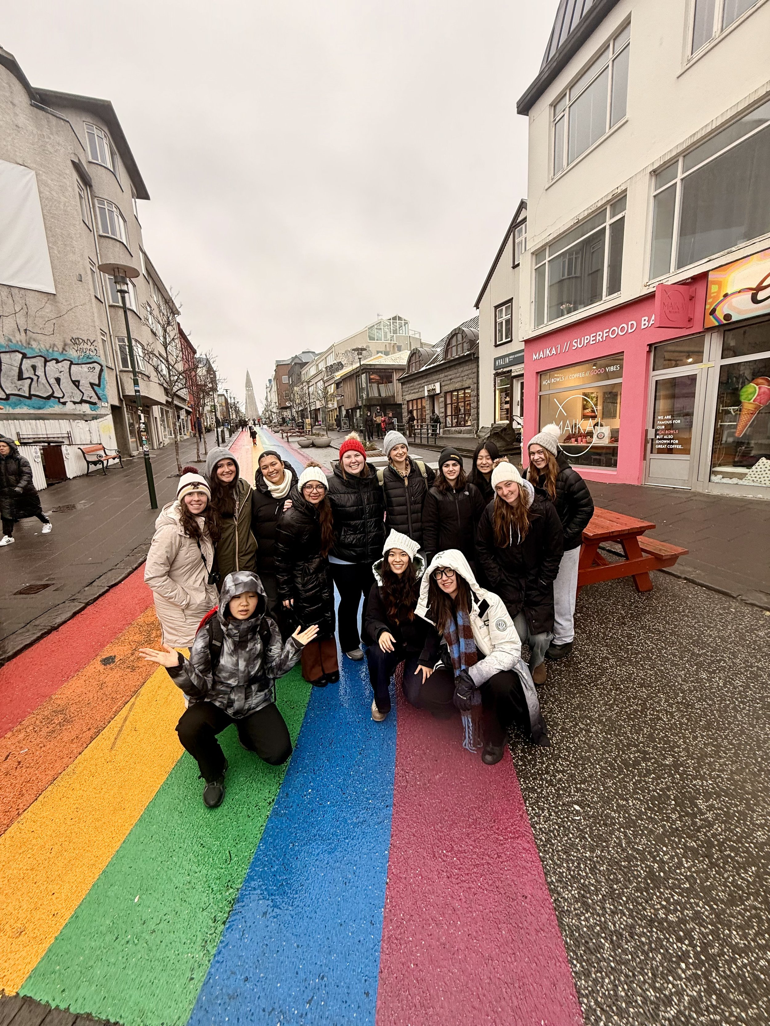 Group of college students standing on Iceland's 'rainbow road', a rainbow-colored crosswalk on a city street, smiling at the camera.