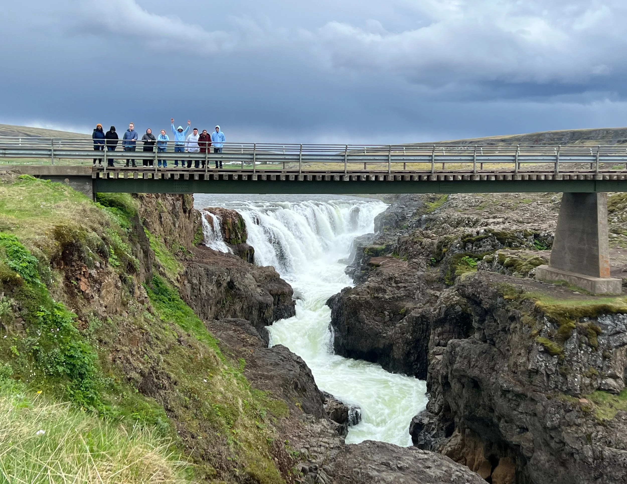 A group of people standing on a bridge over a river, with waterfalls beneath, under a cloudy sky.