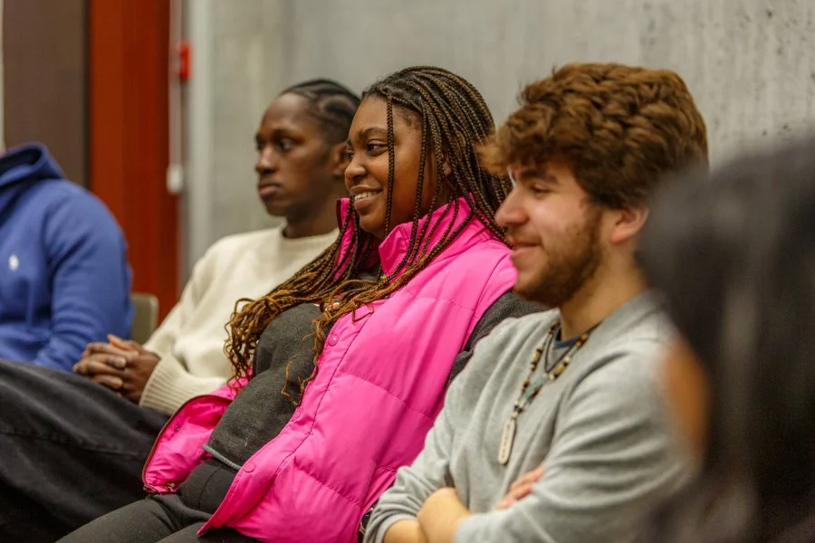 A group of diverse young adults sitting in a room, listening attentively, with one woman smiling and wearing a bright pink vest.