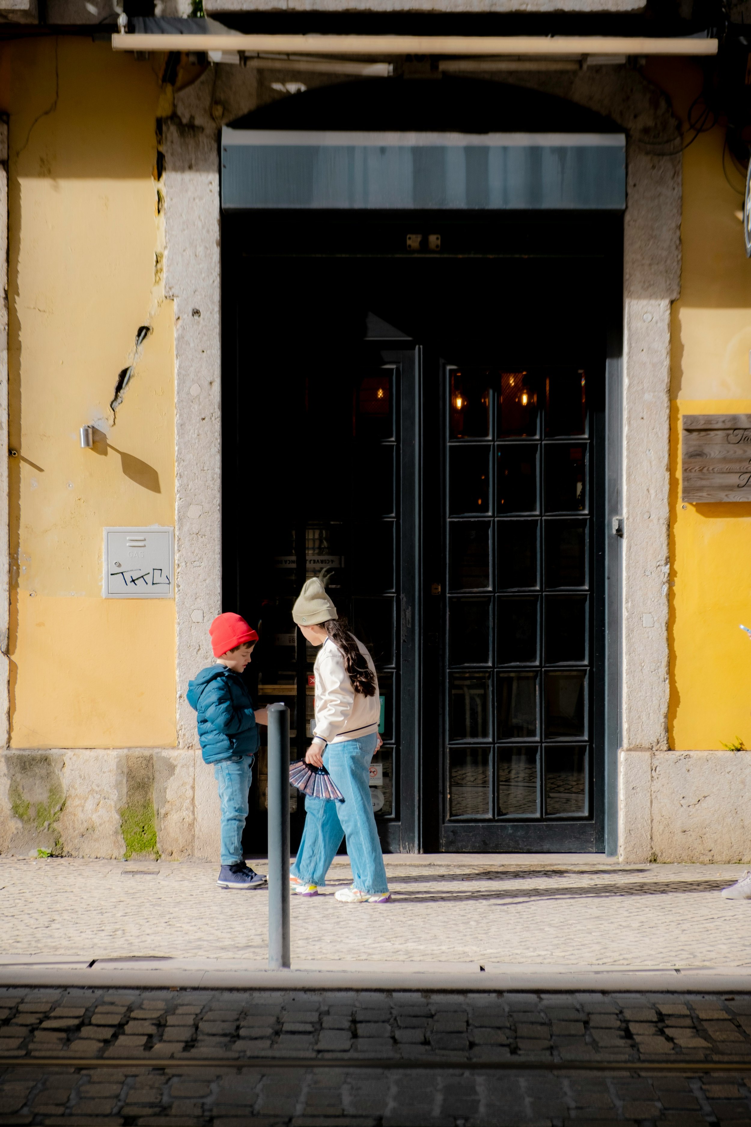 Two children, a boy and a girl, standing outside a building near a black door, engaged in conversation. The girl is holding a fan and wearing a beige beanie, white jacket, and blue jeans. The boy is wearing a red beanie, blue jacket, and jeans. They are standing on a cobblestone street with a yellow wall and a window to the right.