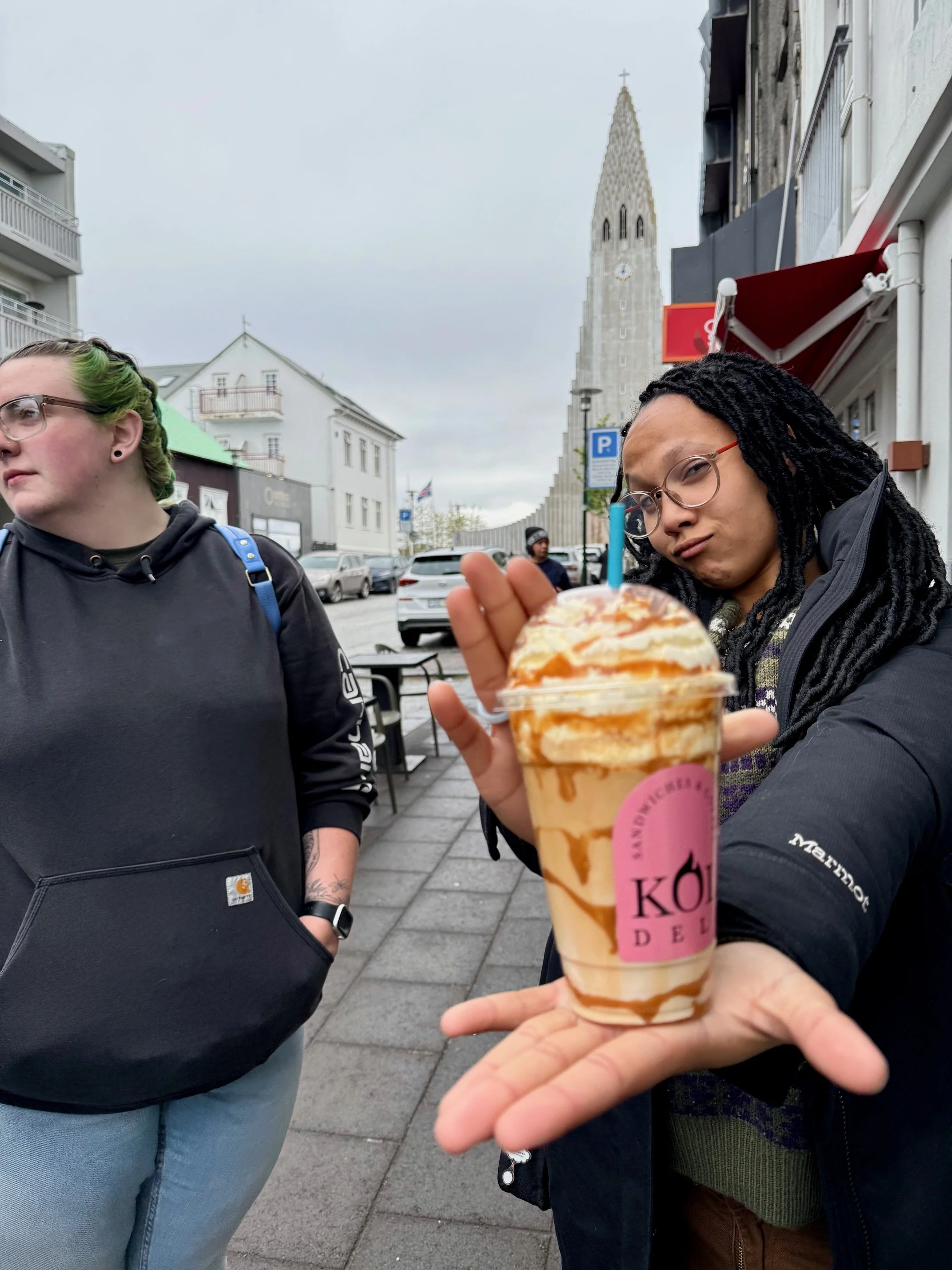 A person with braided hair and glasses holding a caramel drizzle ice cream cone with caramel topping in an outdoor street setting, with two women nearby and historic buildings in the background.