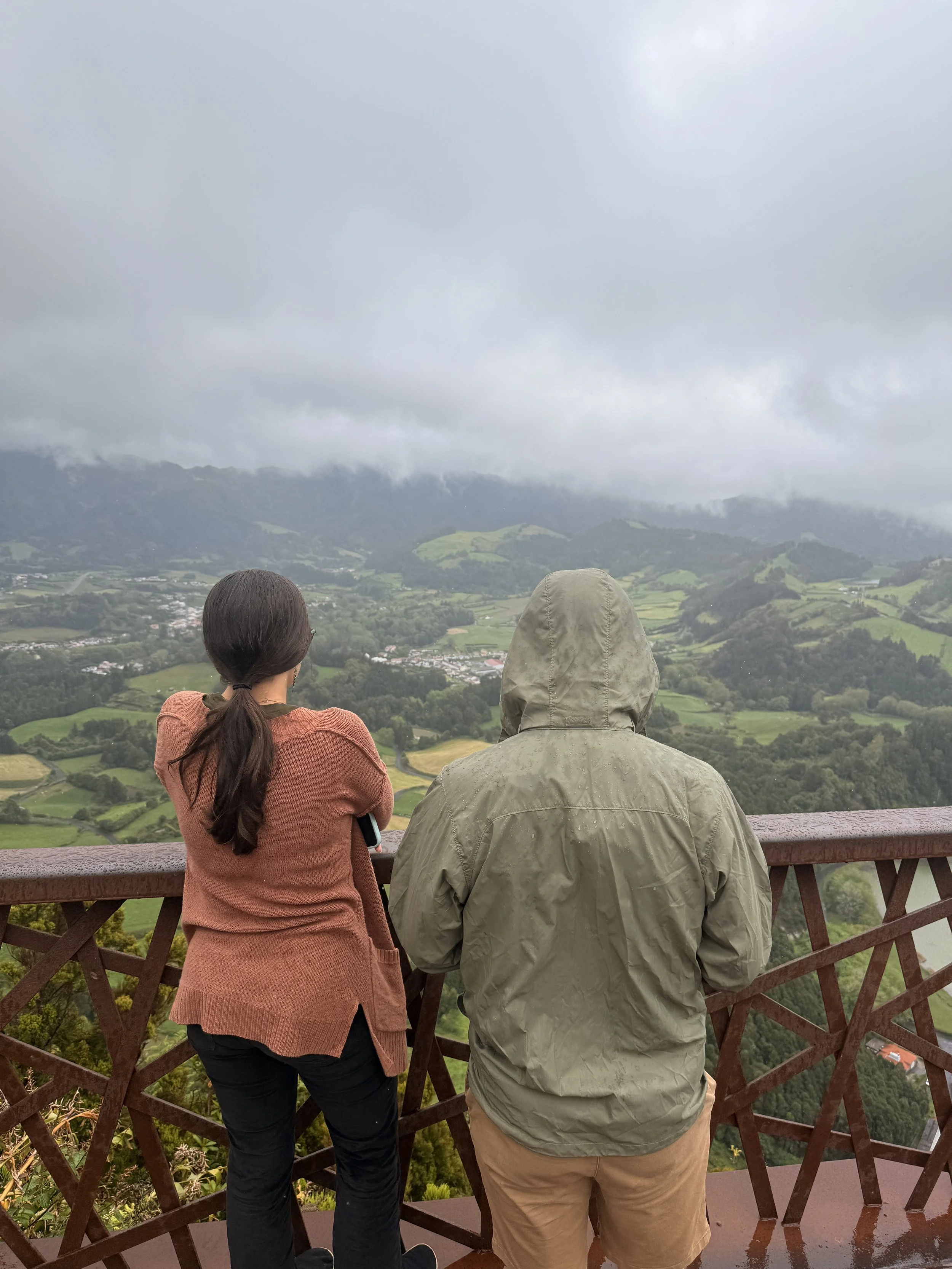 Two people are standing on a viewing platform, looking at a cloudy landscape with green hills and valleys.