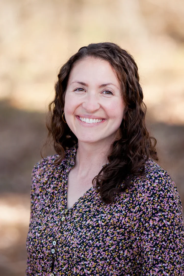 A woman with curly brown hair, wearing a floral blouse, smiling outdoors with a blurred natural background.