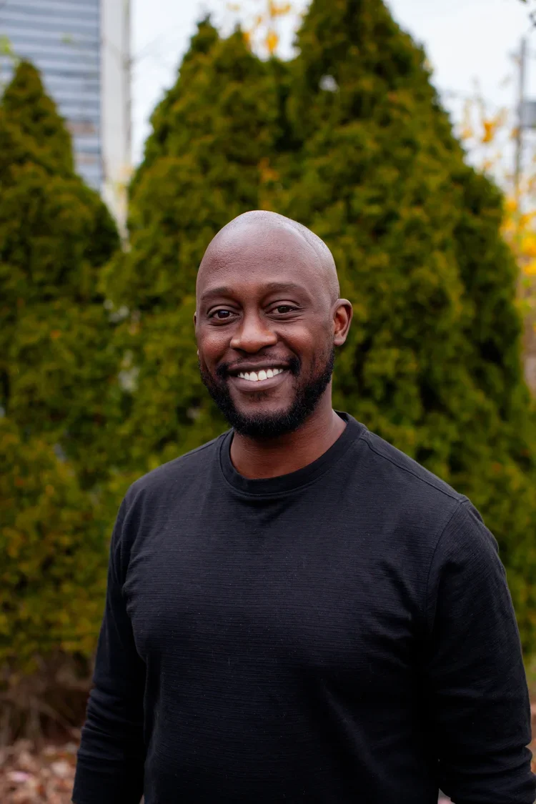 Smiling man with a beard in a black shirt standing outdoors in front of greenery and trees.