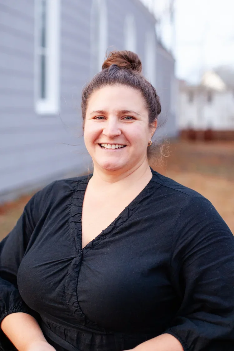 A woman with brown hair in a bun, wearing a black top, standing outdoors in front of a house with gray siding and white windows, smiling.