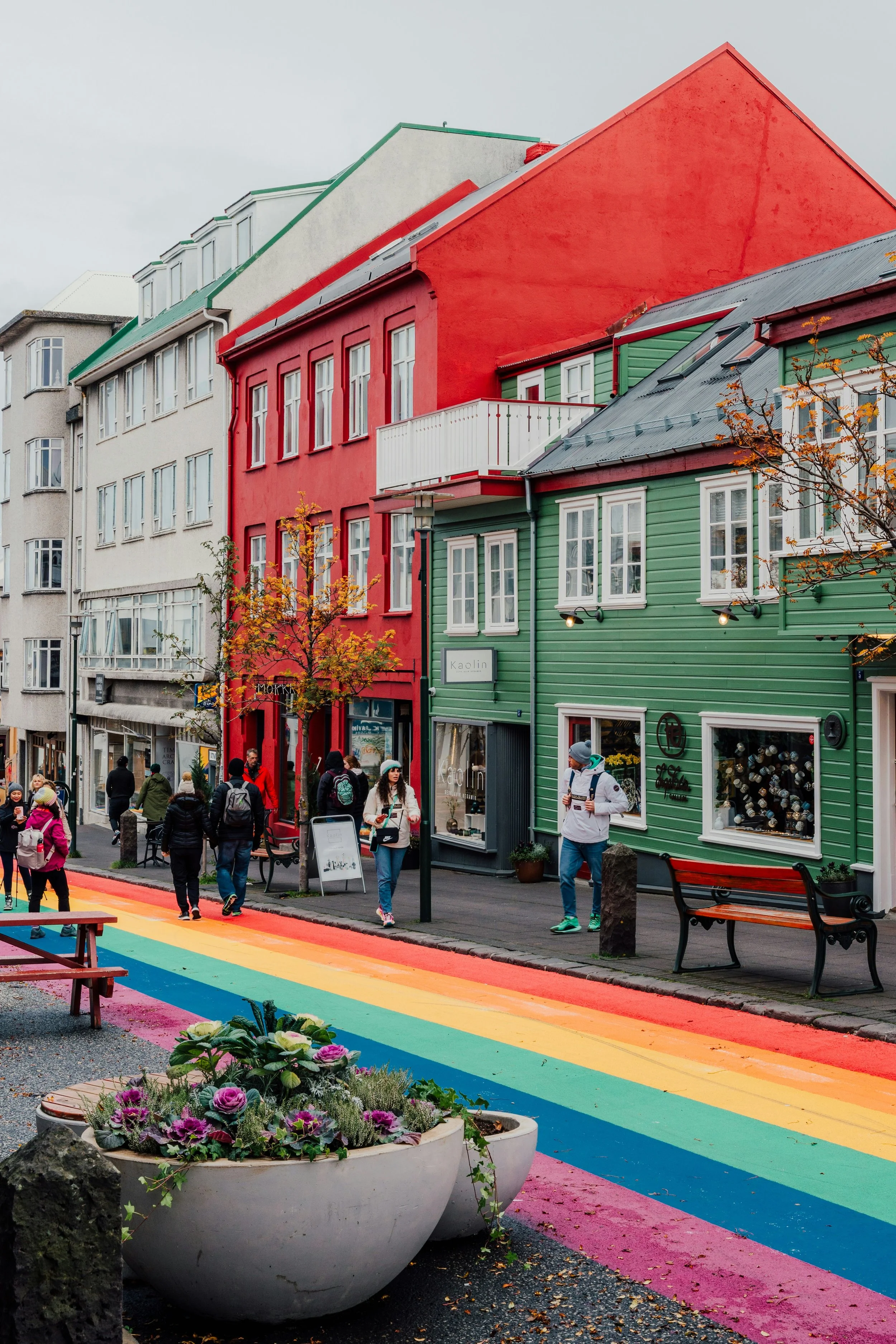 Colorful street scene with rainbow-colored pedestrian crossing, people walking, and vibrant buildings painted in red, green, and white, with flower planters in the foreground.